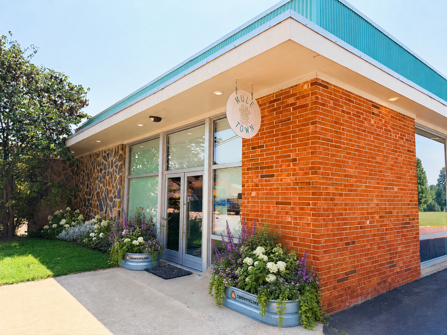 Exterior of a small shop or café with a sign reading "MULE TOWN" featuring a paw print. The building combines red brick and stone siding, with large glass windows and doors. There are flower planters with white and purple flowers on either side of the entrance, and greenery surrounds the building.