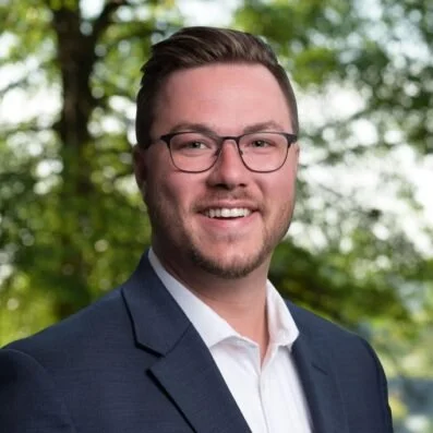 A man in a suit and glasses smiling outdoors with trees in the background.
