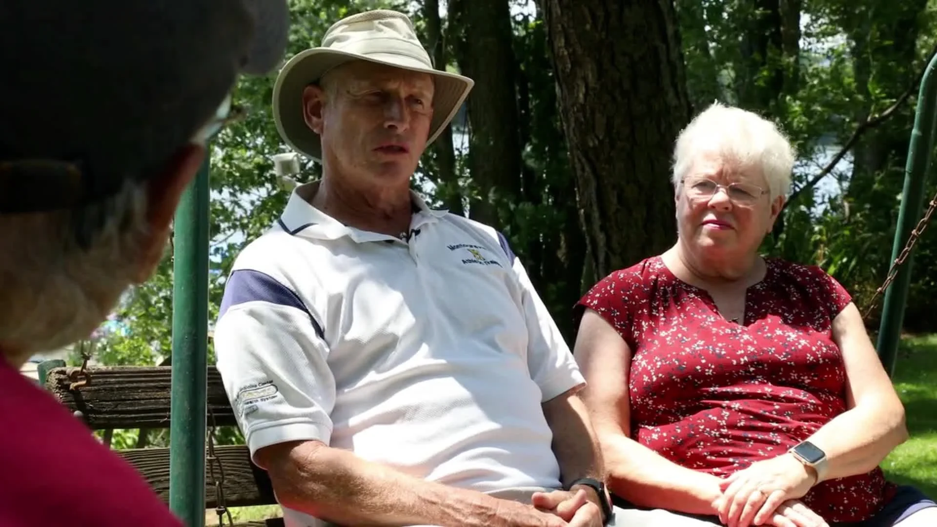 Three elderly people sitting outdoors on a sunny day, engaged in a conversation; a man in a hat and a woman in a red blouse are clearly visible, with another person partially visible in the foreground.