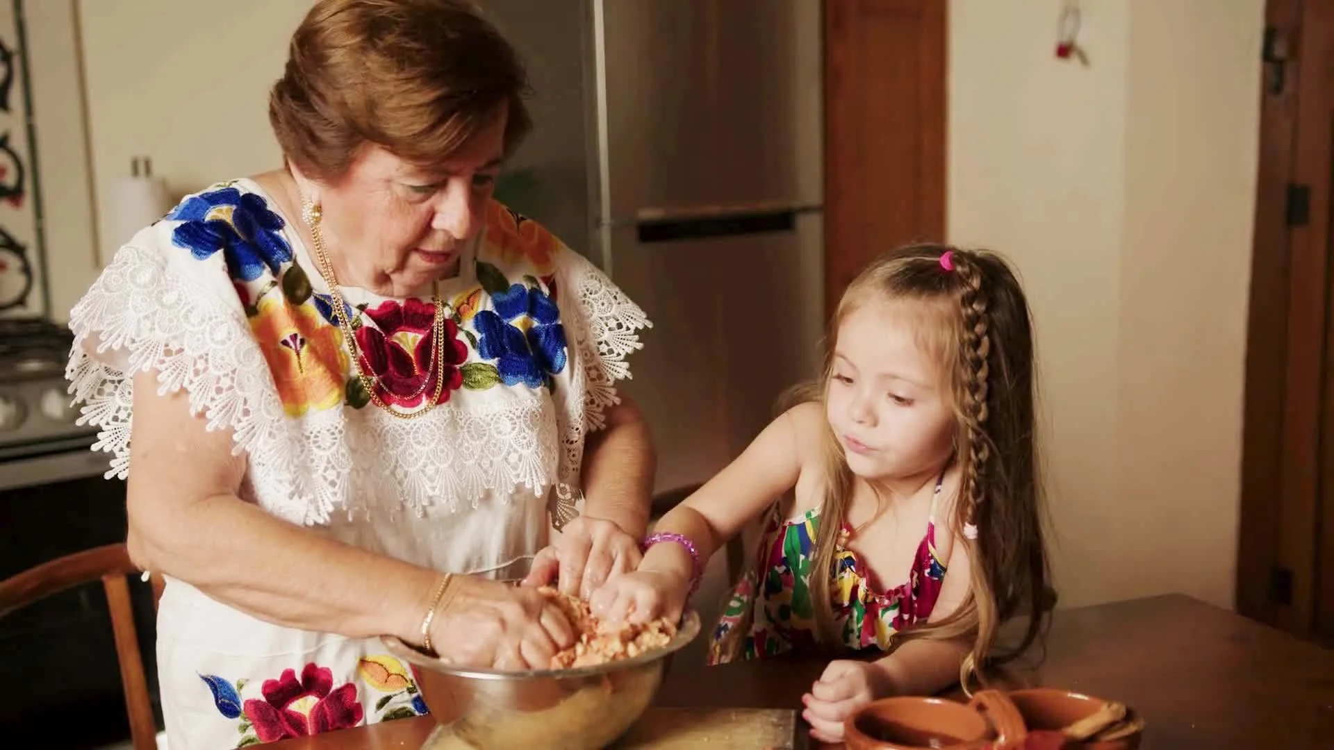 An elderly woman and a young girl are preparing food together in a kitchen, with the woman helping the girl mix ingredients in a large bowl.