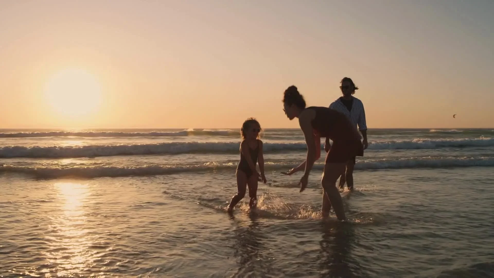 Family enjoying time together at the beach during sunset, with waves in the background and a person in the distance kite surfing.
