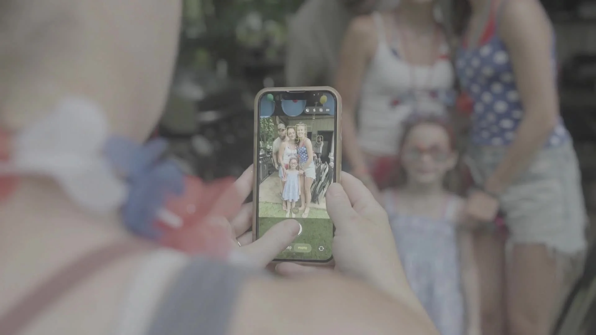 Person taking a photo with a smartphone of a family standing outdoors, with children and adults smiling, while other people are visible in the background.
