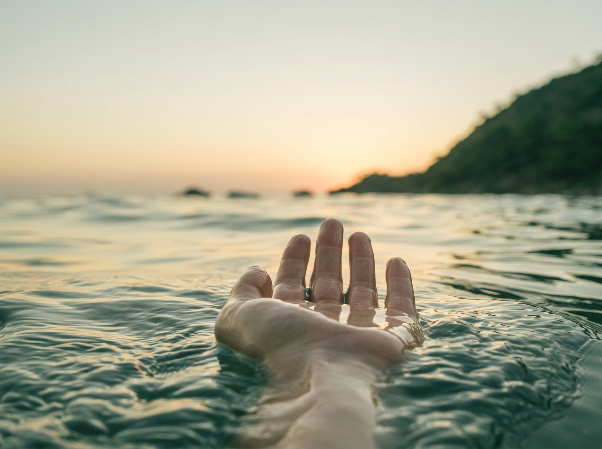 A hand reaching out of water during sunset with a blurred shoreline and hills in the background.