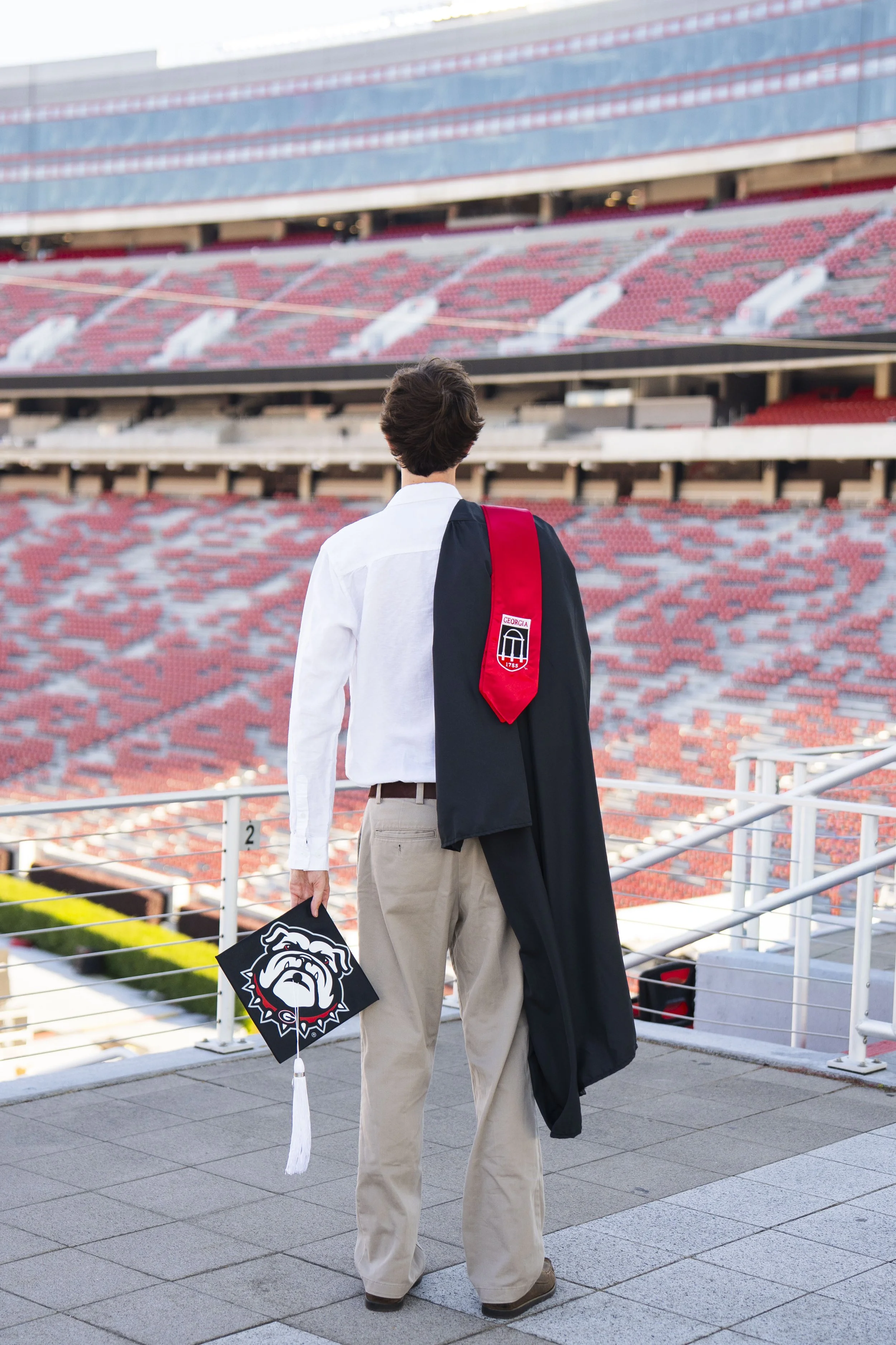 A graduate standing in Sanford stadium with empty red seats, wearing a black gown and holding a Bulldogs themed graduation cap, with a young man in a white shirt and beige pants nearby.