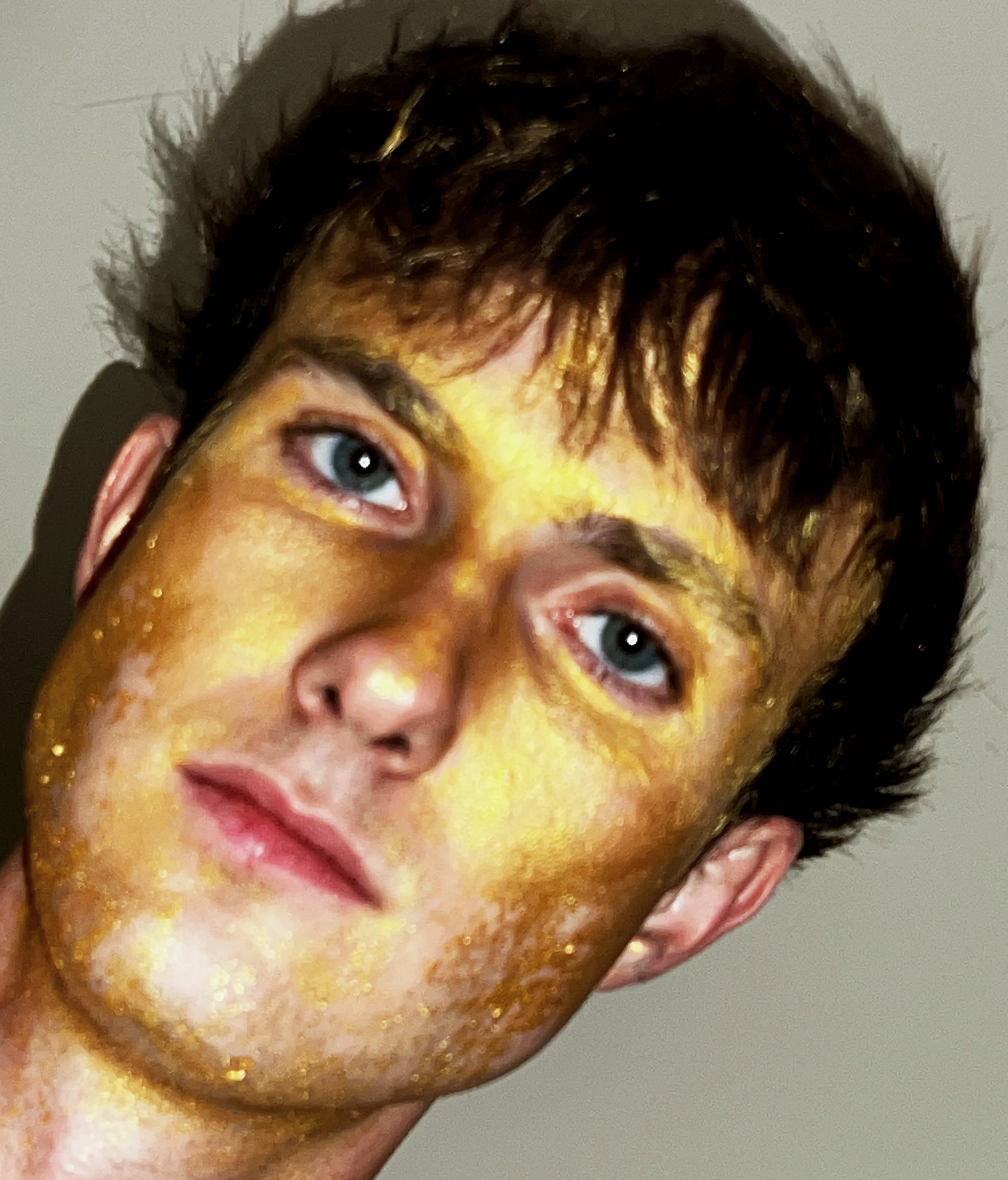Close-up of a person's face with a yellow painted face and glitter on skin, featuring blue eyes, dark, tousled hair, and an ear visible on the right side.
