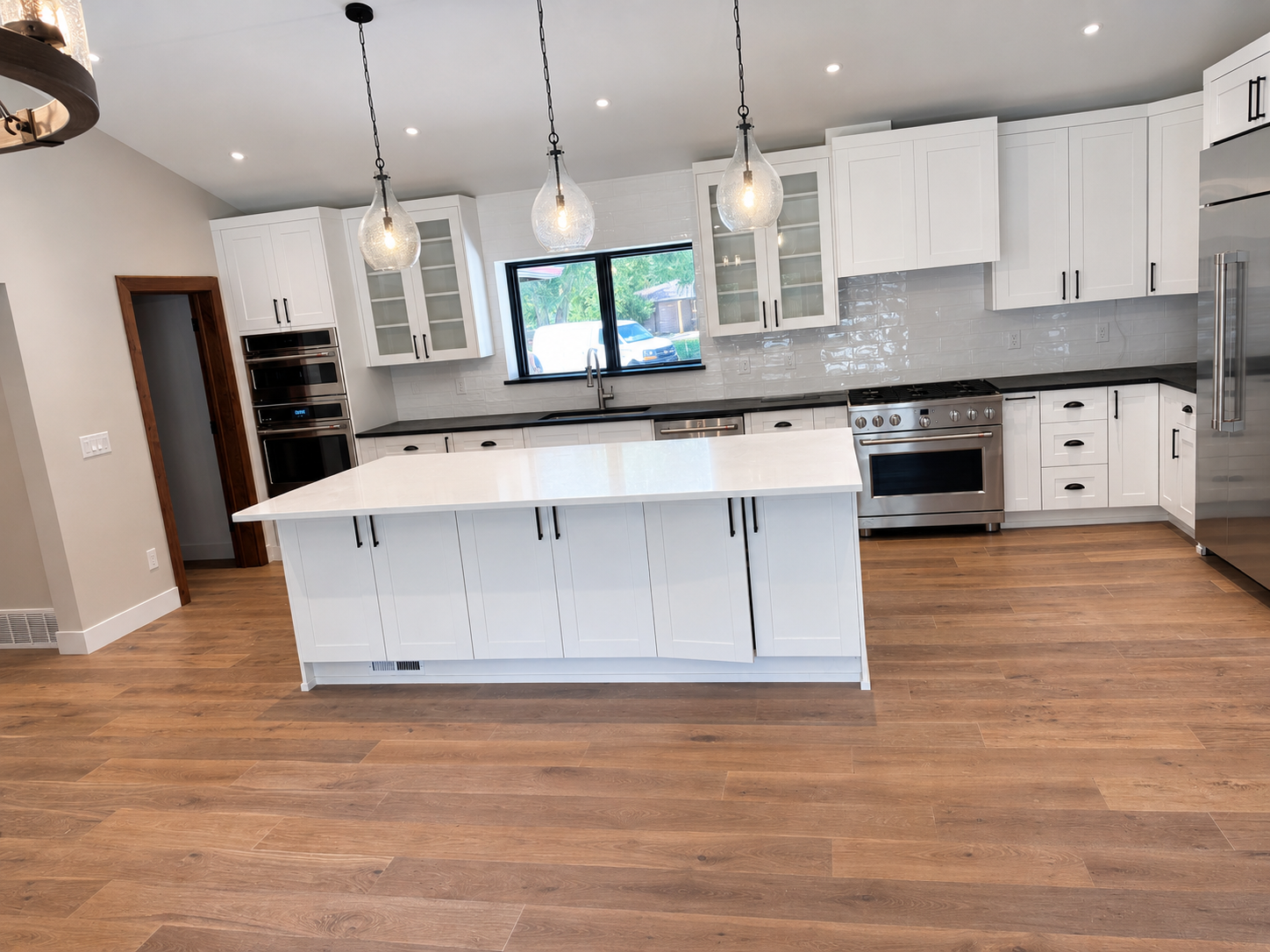 Modern kitchen with white cabinets, black countertops, stainless steel appliances, wooden floors, and a central island with a white countertop, pendant lights above, and a window over the sink.