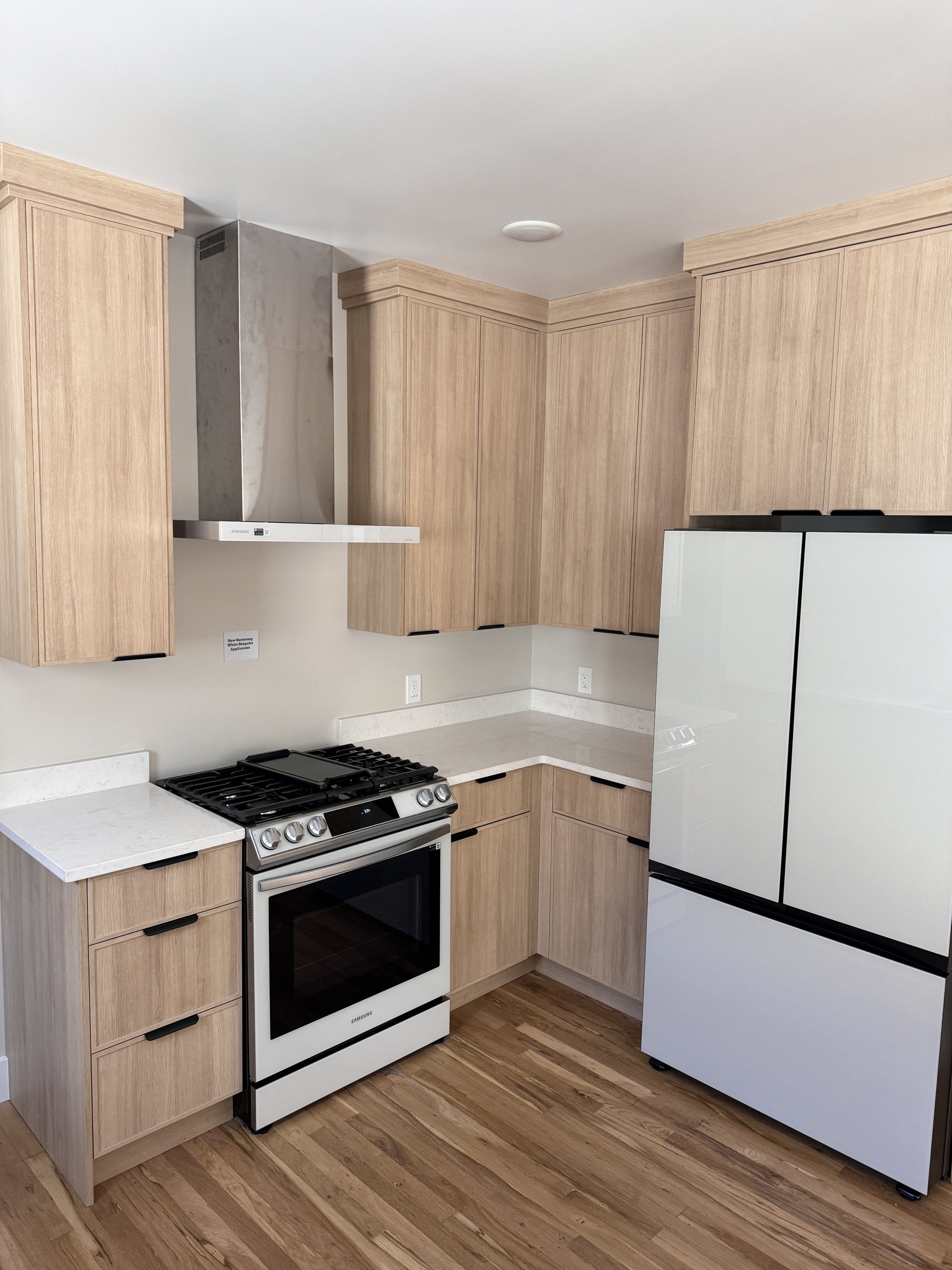 A modern kitchen with light wood cabinets, a white oven, a stainless steel range hood, a white refrigerator, and hardwood flooring.