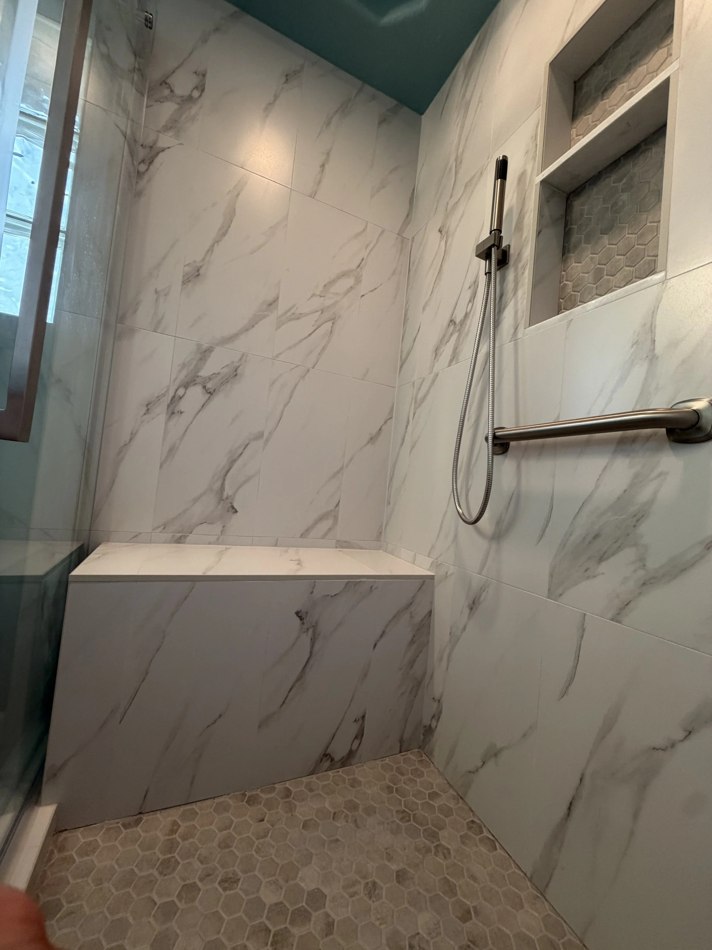 A walk-in shower with large white marble tiles with gray veining on the walls, a rectangular built-in niche with gray hexagonal tiles, a metal showerhead with a hose, and a grab bar. The floor has small beige hexagonal tiles.