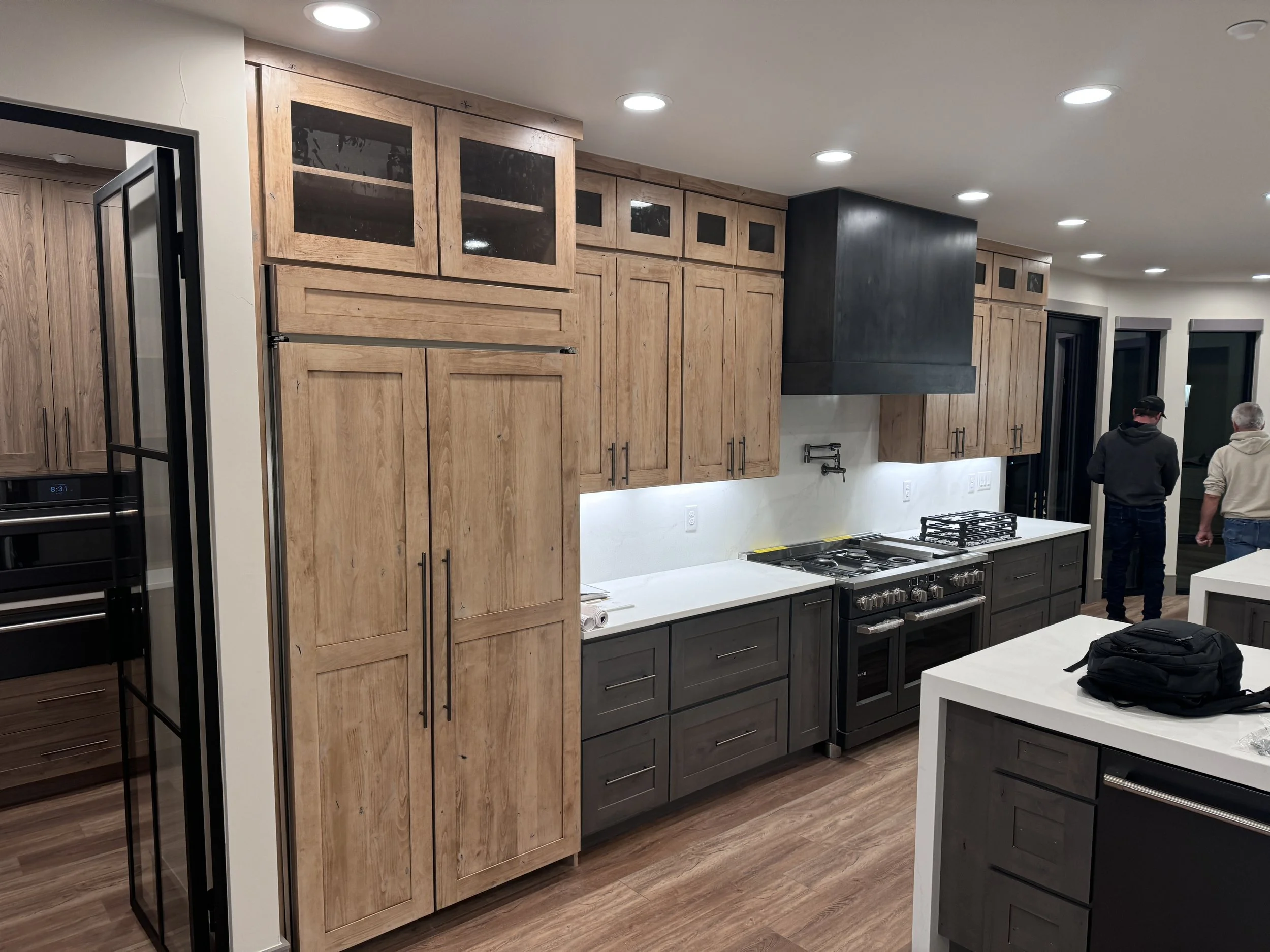 Modern kitchen with wooden cabinets, a stainless steel stove, and a white countertop. There are recessed ceiling lights and two men standing near a door on the right side.