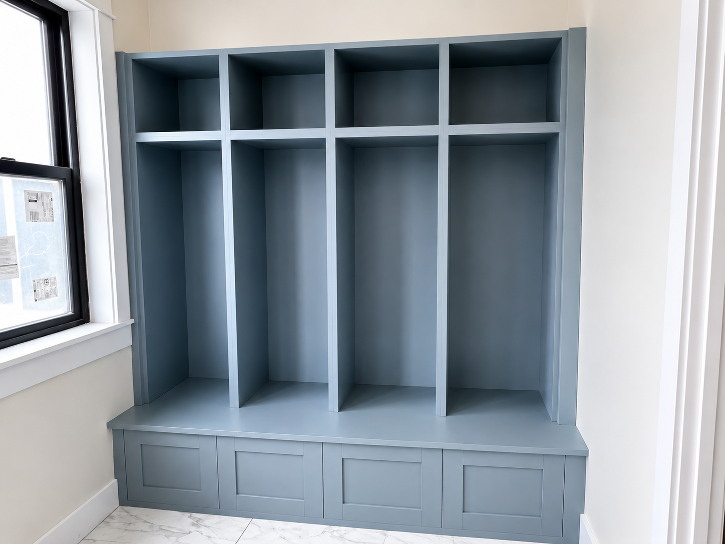 A blue built-in storage unit with shelves and cabinets against a white wall, next to a window with black frame.