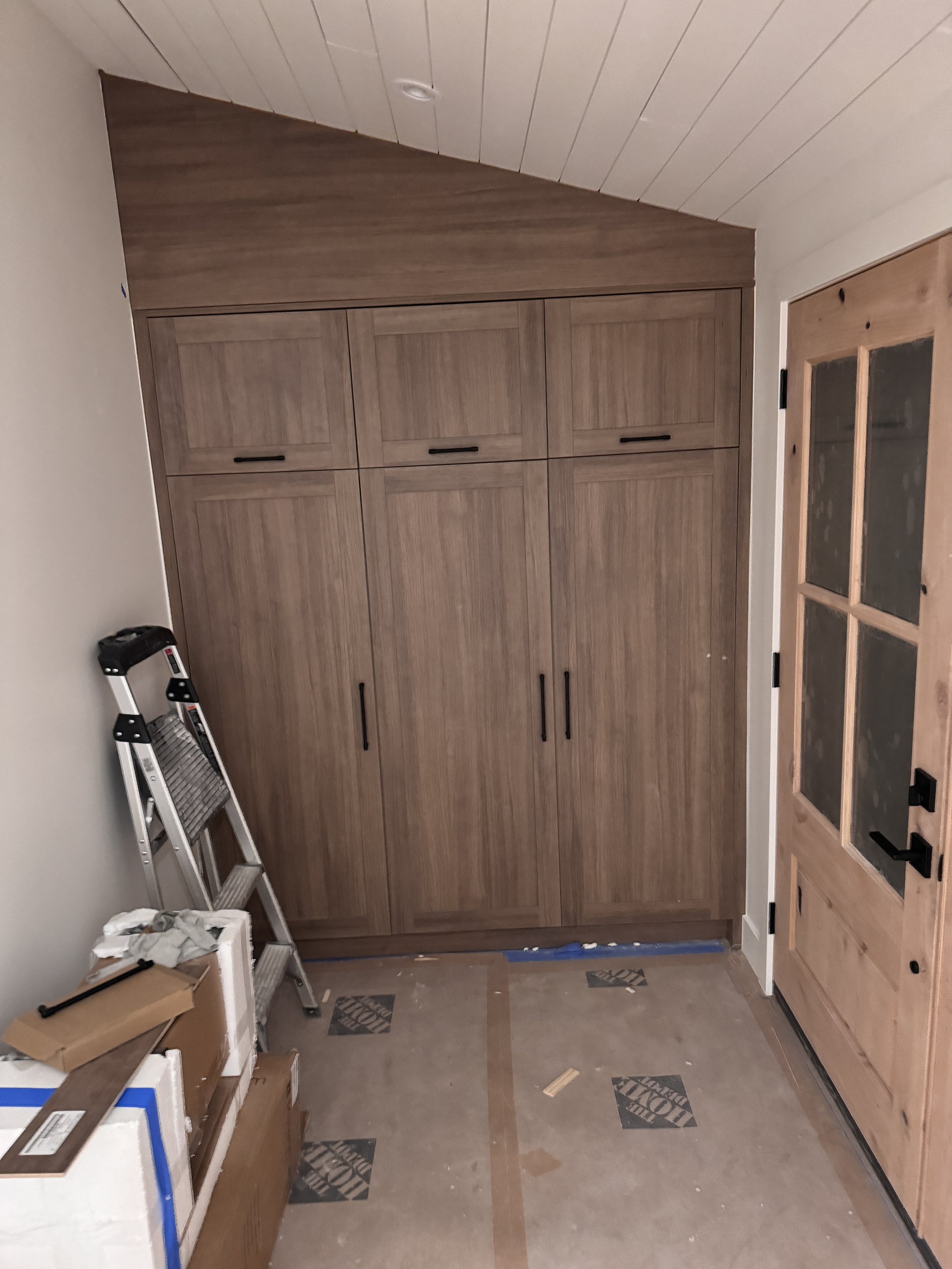 A room under construction with wooden cabinets, a step ladder, boxes, a cardboard box, and a plastic container on the floor, and a door with glass panels.
