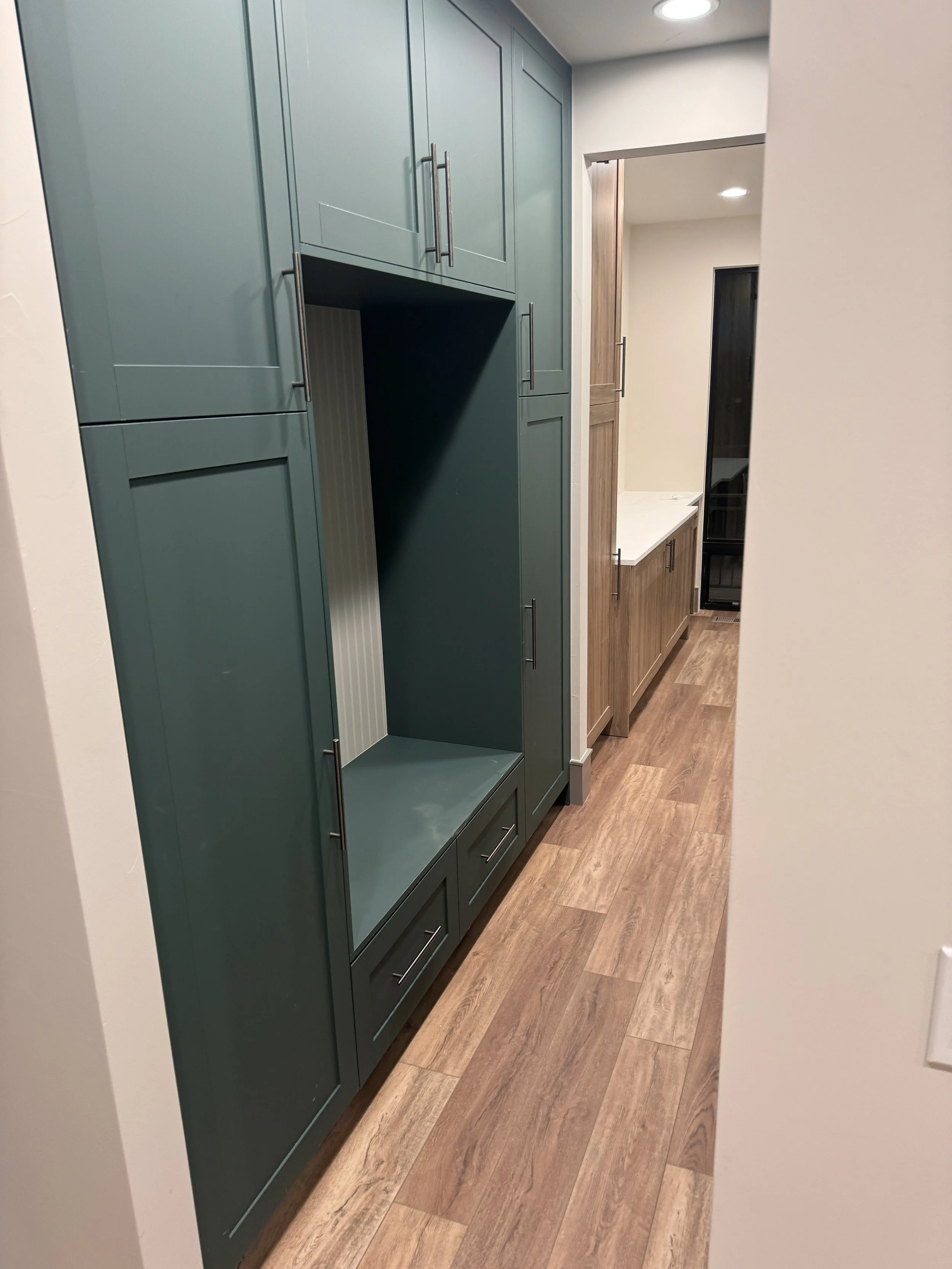 A hallway with a green tall cabinet on the left side, a wooden cabinet with drawers in the background, and a wooden floor.
