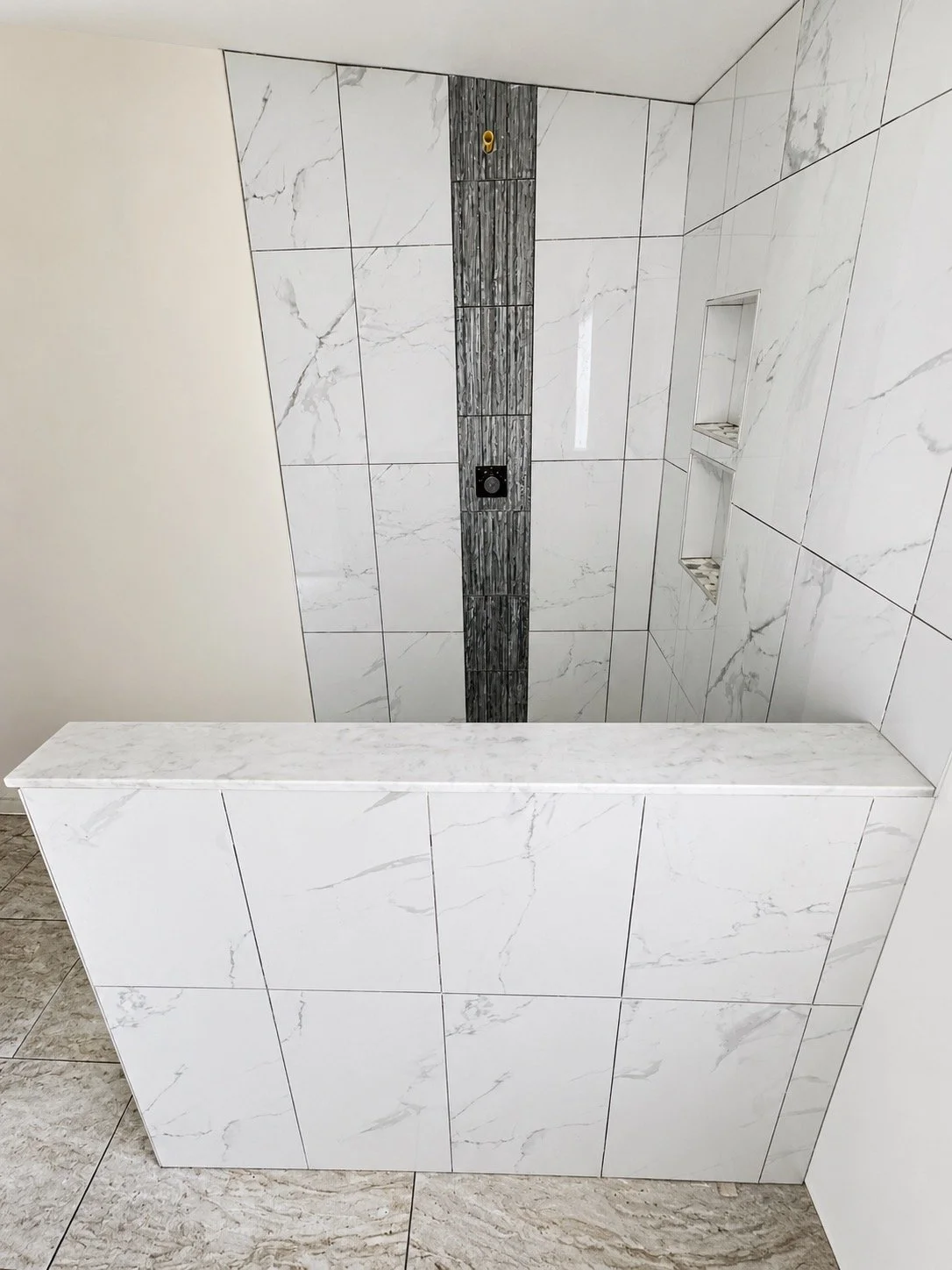 Bathroom shower area with white marble tiles, a dark vertical accent strip with a shower head, built-in niches, and a marble half-wall.