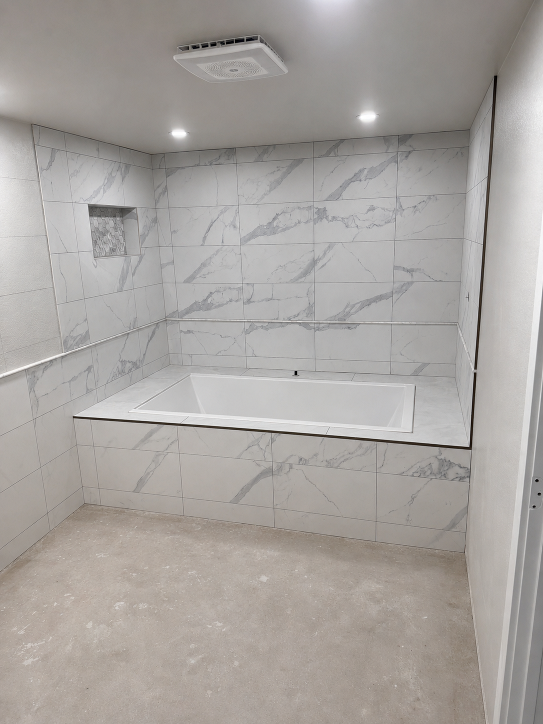 A bathroom with marble tile walls, a built-in bathtub, a recessed shower niche, a ceiling vent, and recessed ceiling lights.