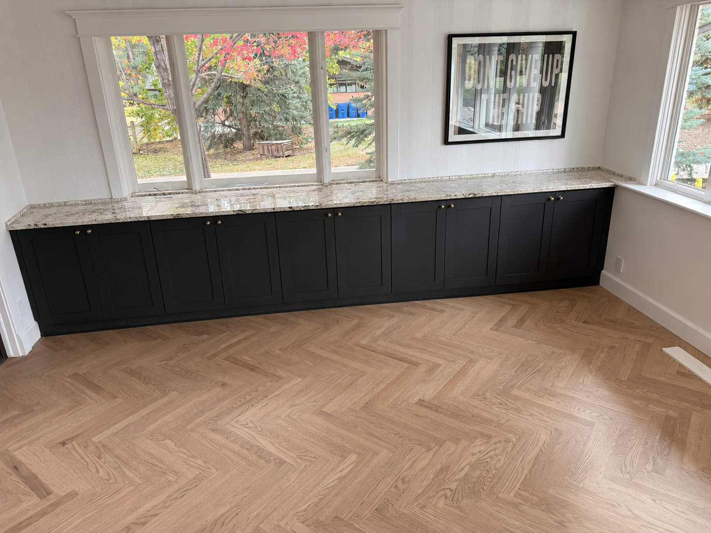 A room with a large window showing trees outside with red and green leaves. There is a black cabinet with a granite countertop below the window. A framed poster on the wall reads 'DON'T GIVE UP ON HOPE'. The room has light-colored hardwood floors arr