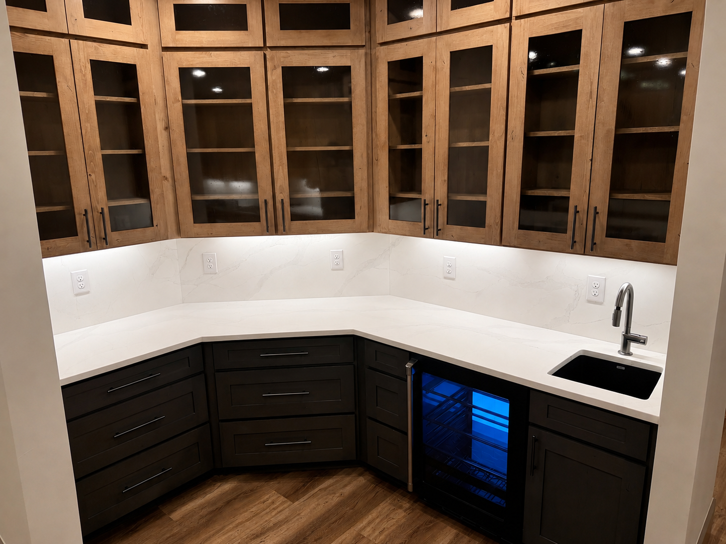 Kitchen corner with wooden upper cabinets, dark lower cabinets, a white marble countertop, a small under-counter wine cooler with blue interior light, and a black sink with a modern faucet.