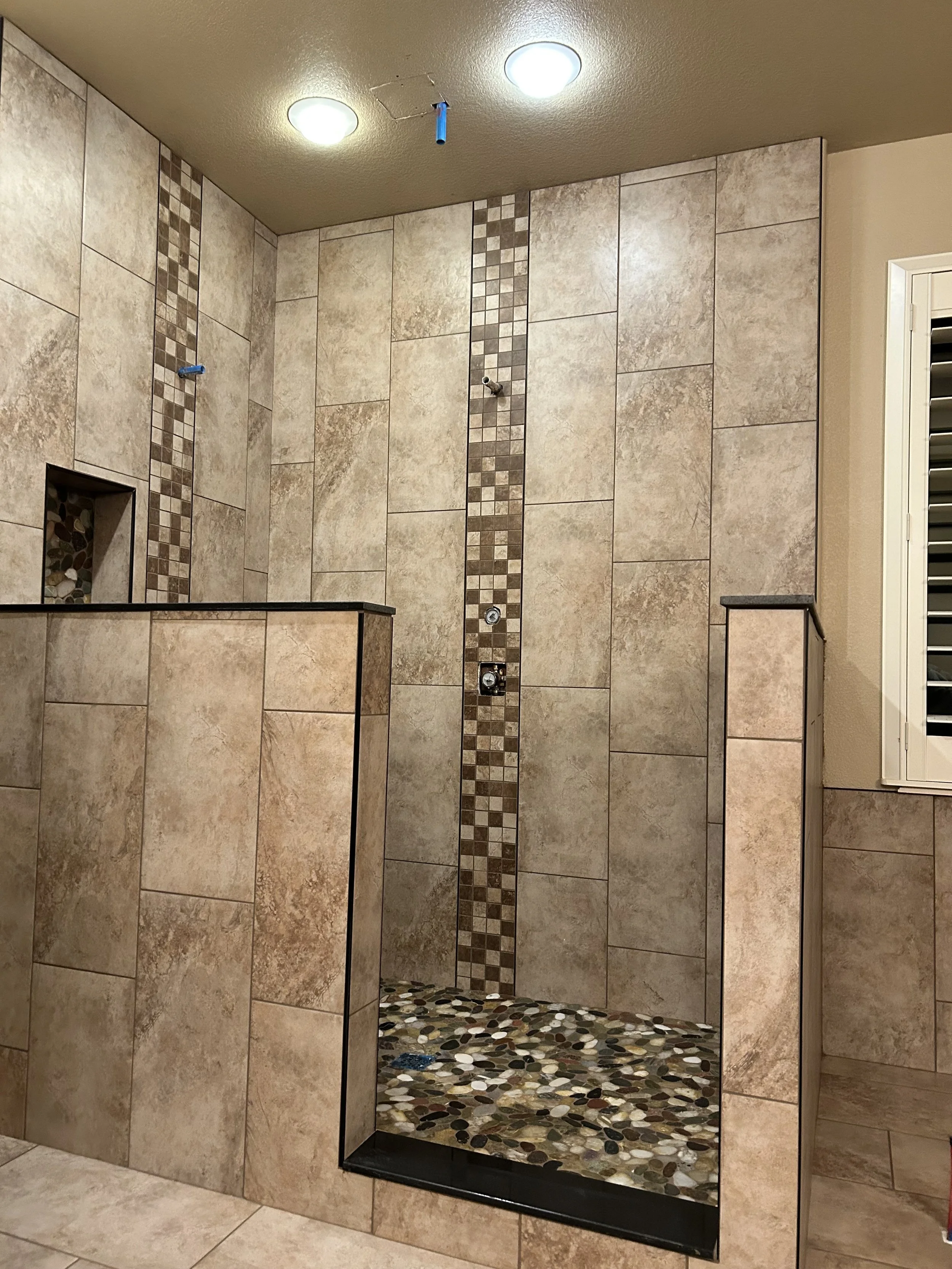 A bathroom walk-in shower with beige tiles, a vertical strip of small square tiling, a pebble stone floor, and a bench on the right side.