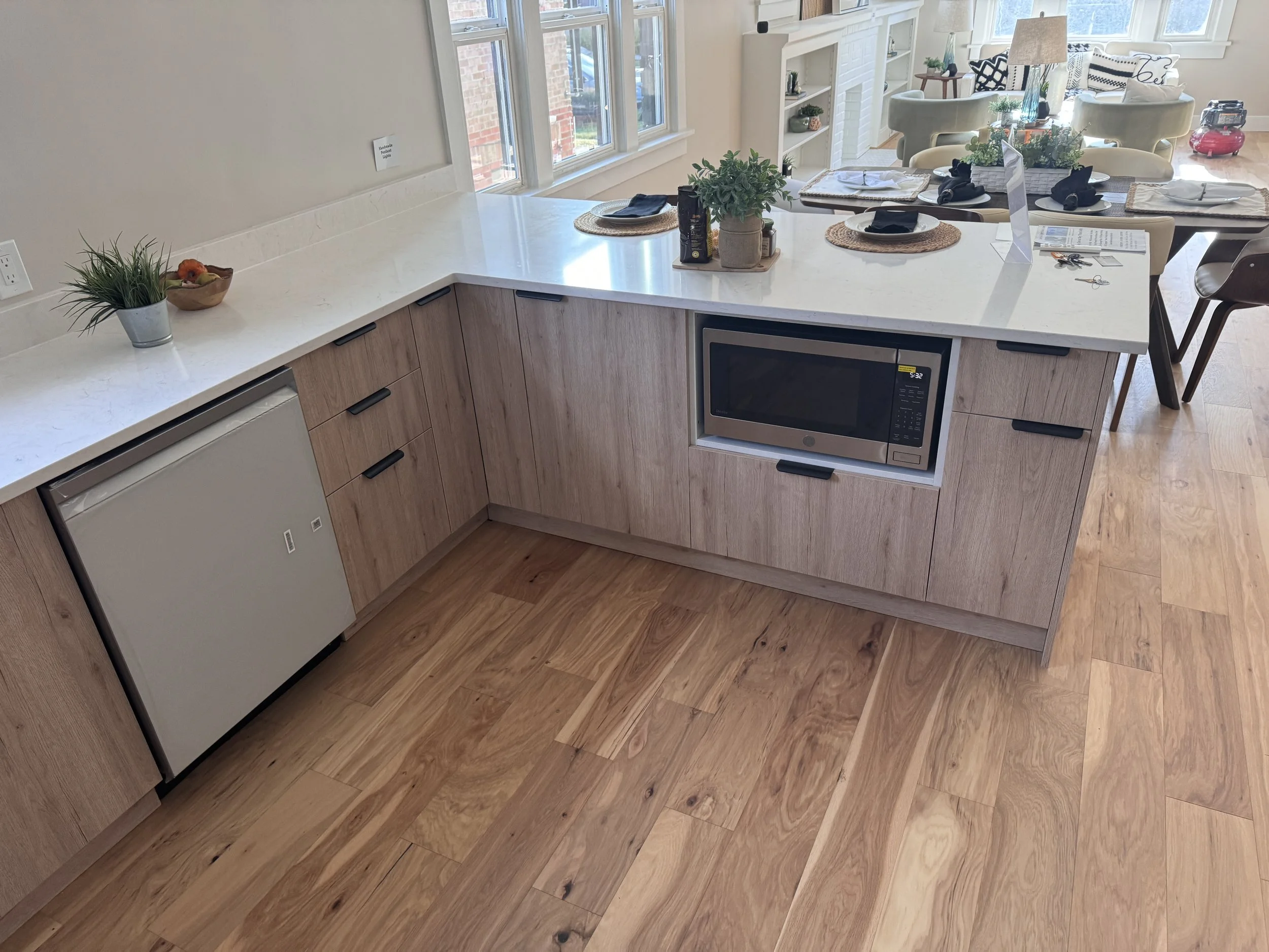 View of a modern kitchen island with a white marble countertop, wooden cabinetry, a microwave oven, and decorative plants, adjacent to a dining area with table settings and natural light from multiple windows.