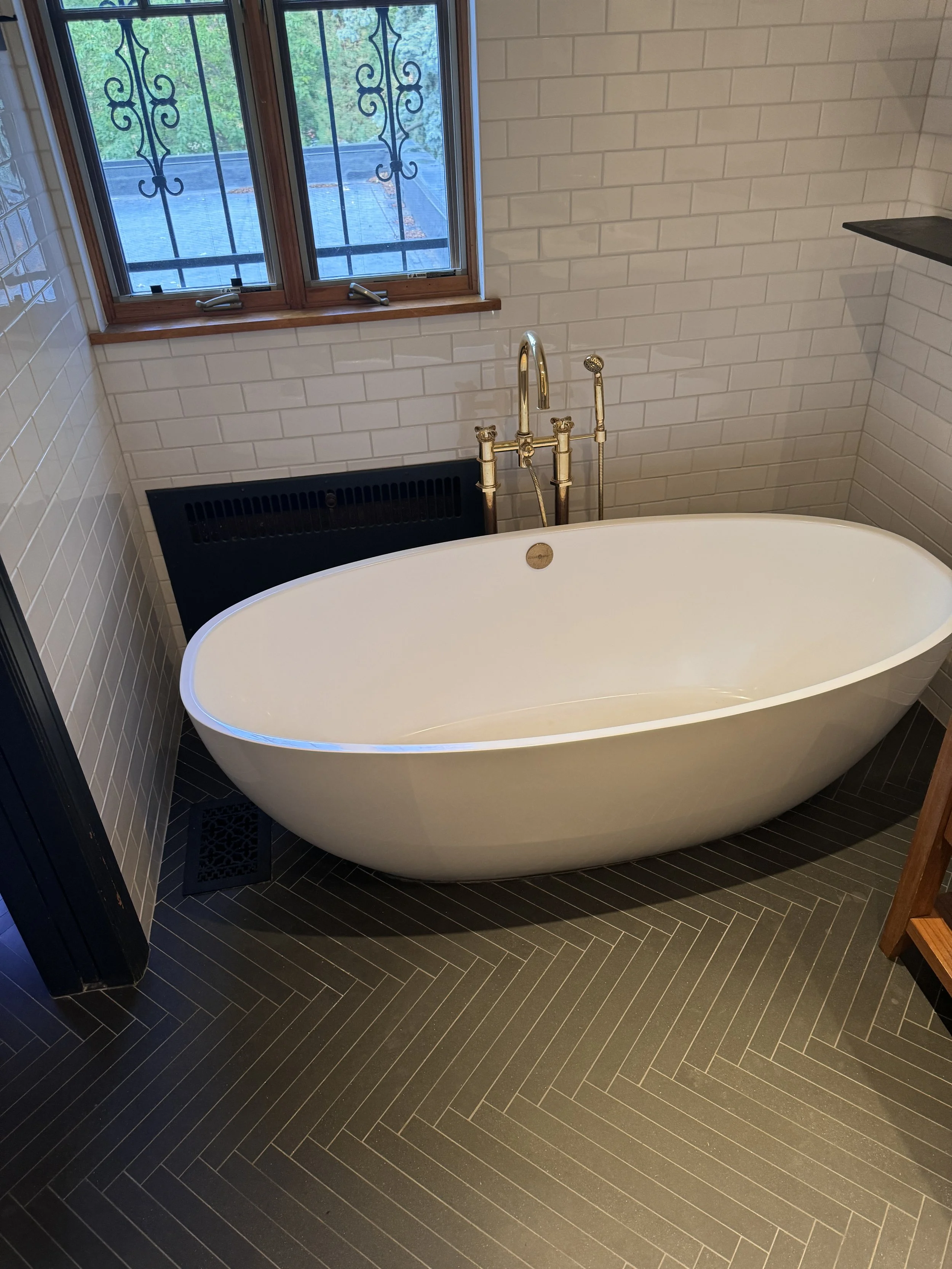 A white freestanding bathtub with a gold faucet and handheld showerhead, next to a window with black wrought iron security bars, in a bathroom with white brick walls and black herringbone patterned floor tiles.