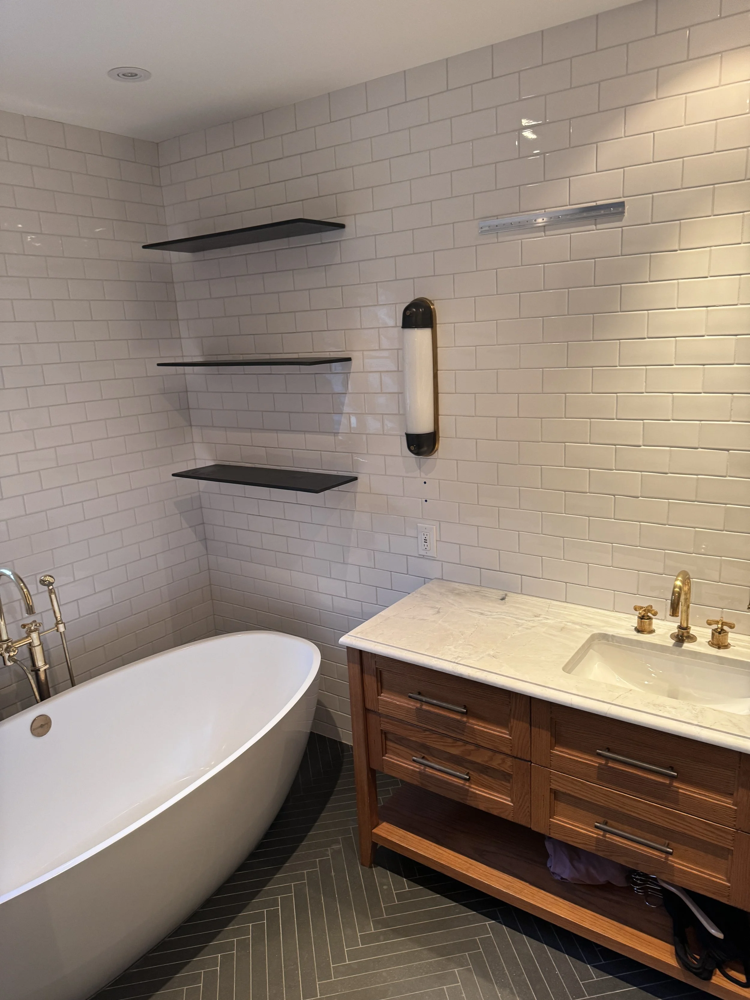 A bathroom with a white bathtub, white brick wall, black corner shelves, wooden vanity with marble countertop, small sink, brass faucet, and a wall-mounted light.