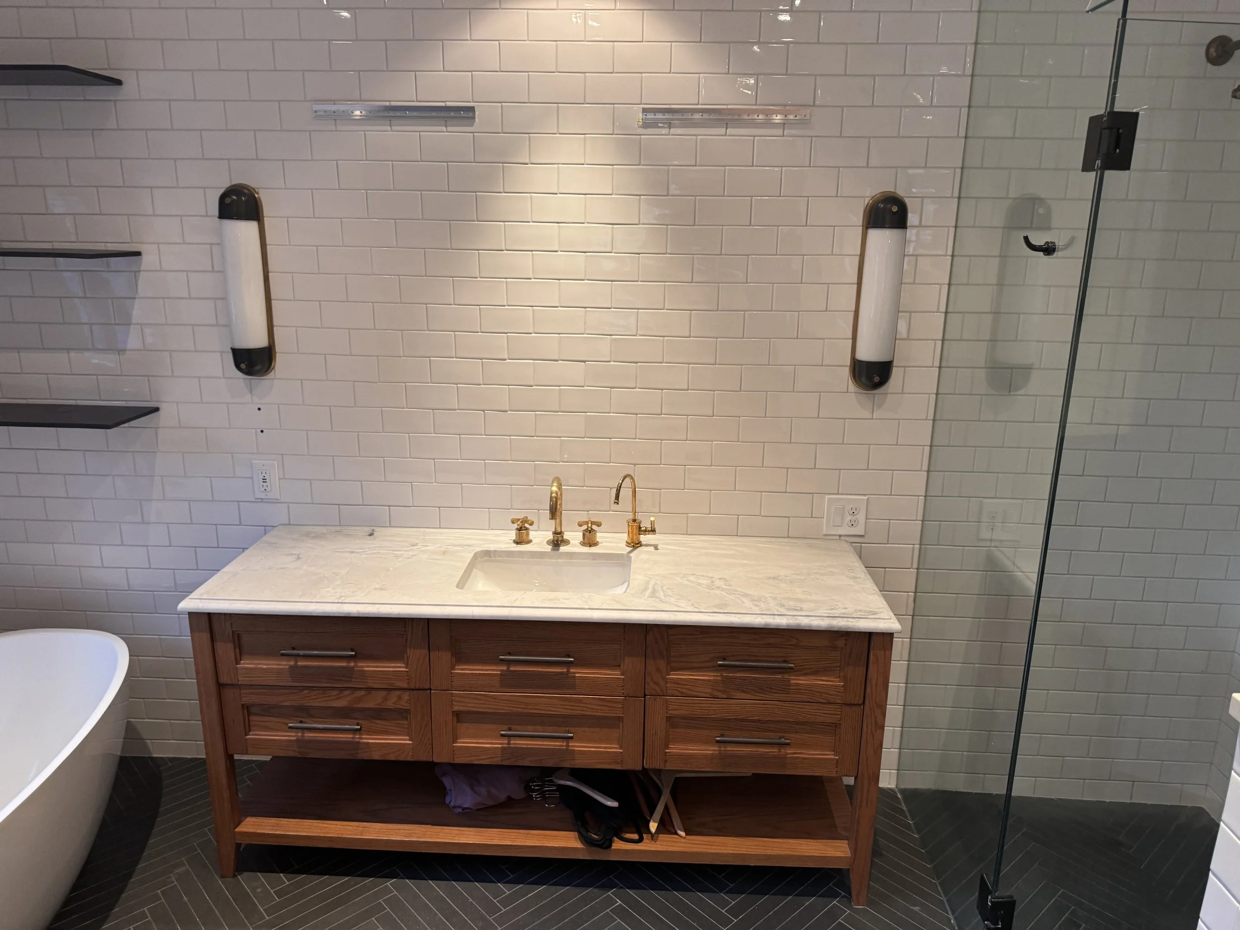 Bathroom sink with a wooden vanity, gold faucet, white marble countertop, white subway tile wall, black wall-mounted towel bars, mirror, black floating shelves, glass shower enclosure, electrical outlet, and part of a bathtub.