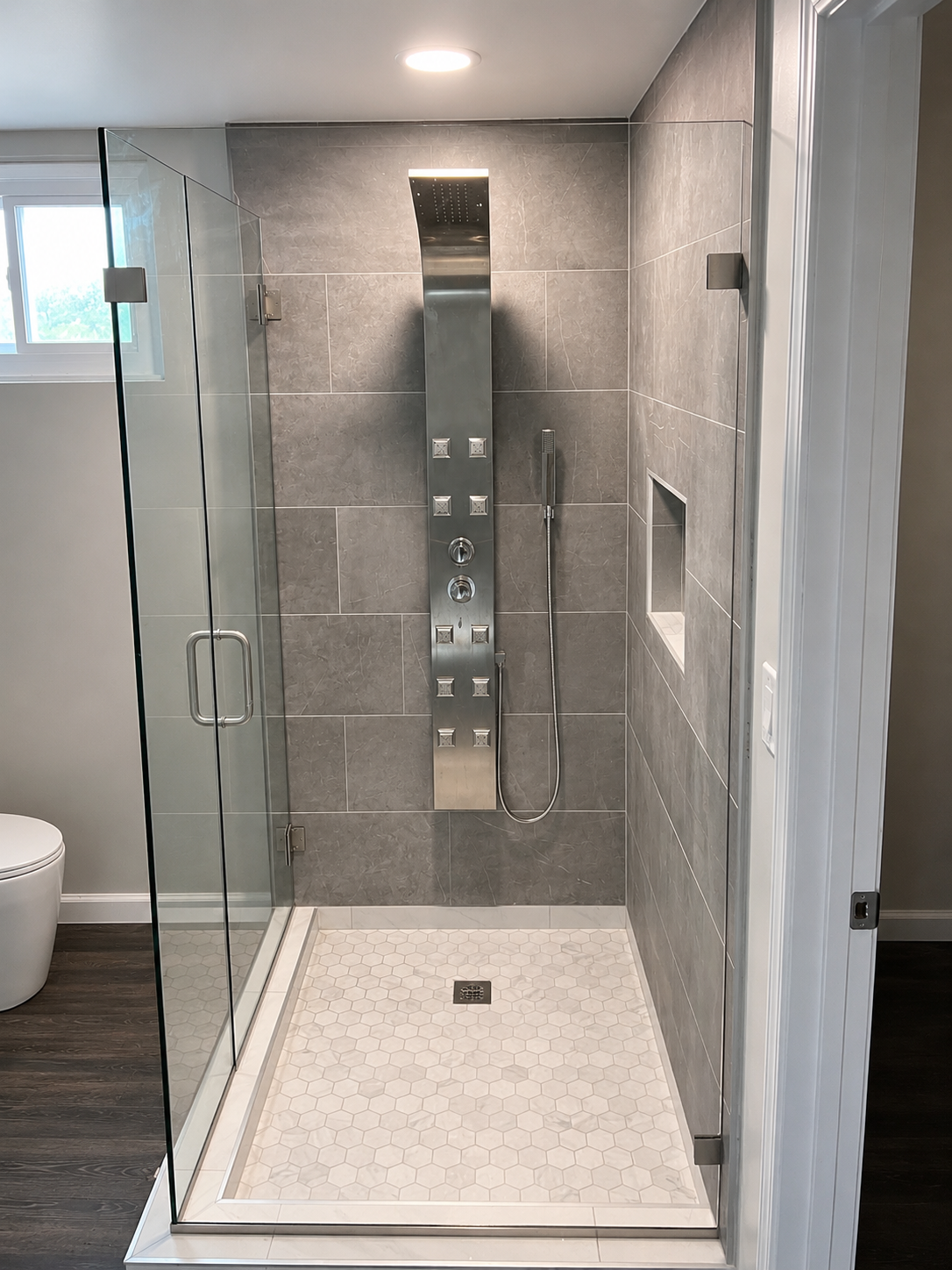 Modern shower with gray tiled walls, a built-in niche, a stainless steel shower panel, a handheld showerhead, and a clear glass enclosure.