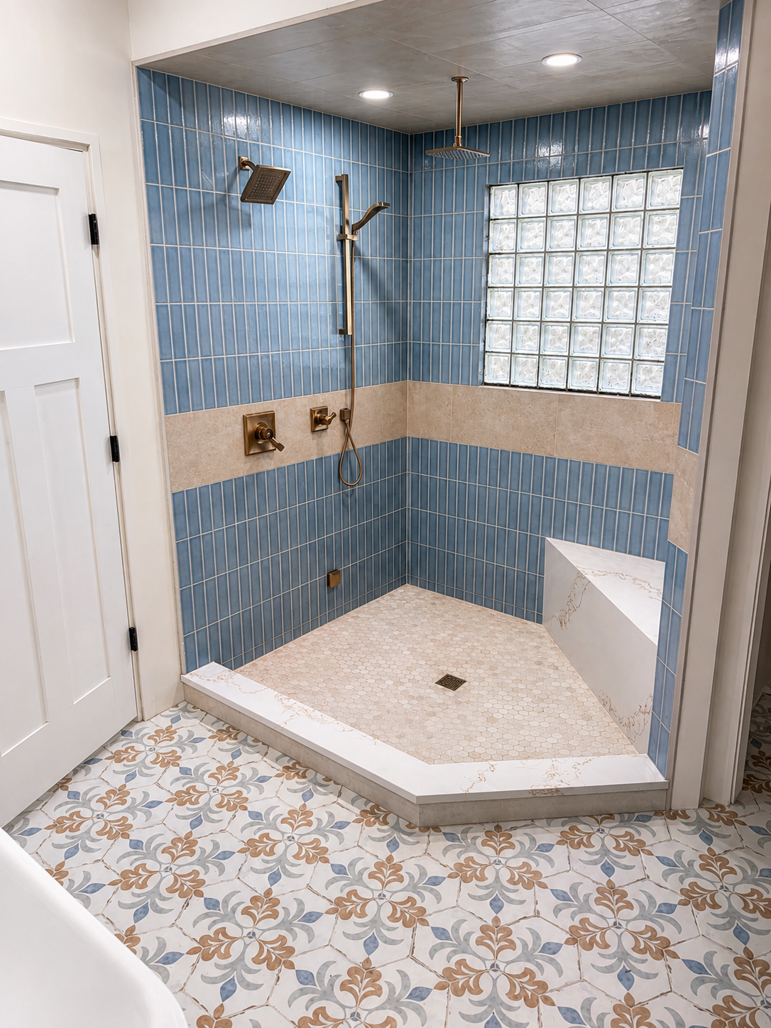 A walk-in shower with blue and beige tiles, a glass block window, and bronze fixtures. The shower has two showerheads and a small built-in bench, with a patterned tile floor outside.