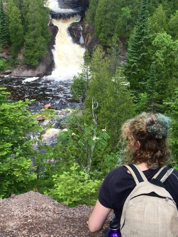 A woman with curly hair and a backpack sitting on a dirt outcrop, looking at a waterfall in a lush green forest of Northshore Minnesota.