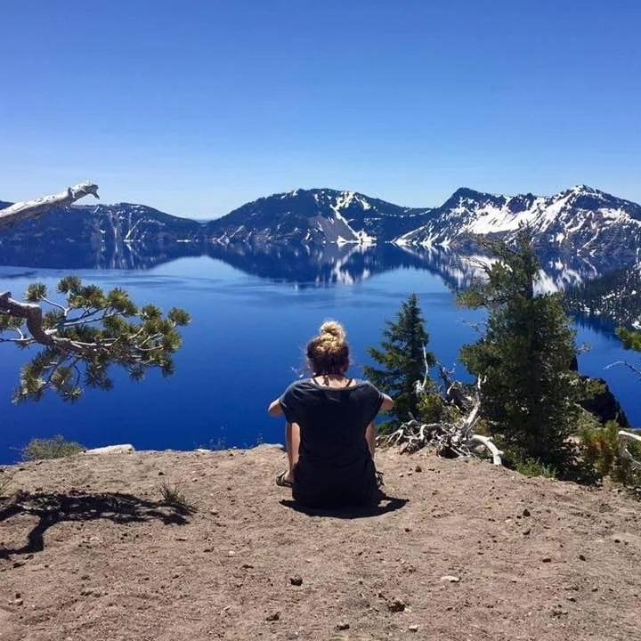 A person sitting on dirt ground overlooking a large blue lake surrounded by snow-capped mountains and pine trees at Crater Lake.