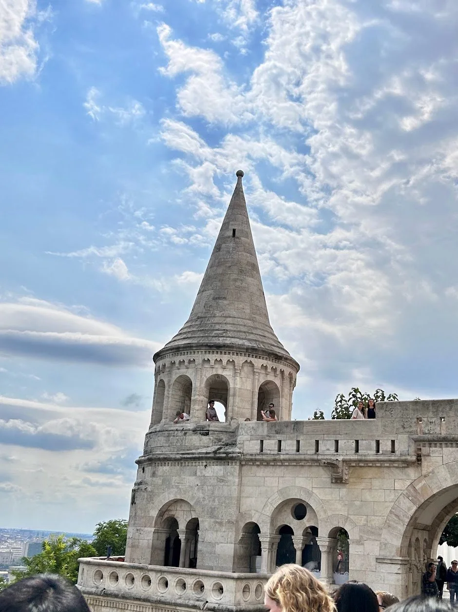 A historic stone tower, Fisherman's Bastion, in Budapest, with a tall, pointed tower and arched windows, set against a partly cloudy sky, with tourists exploring the site.