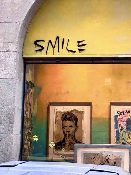 A storefront window with the word 'SMILE' spray-painted above. Inside the window, there are framed artworks, including a portrait of a person with their finger to their lips, and various other colorful pieces. Barcelona Spain.