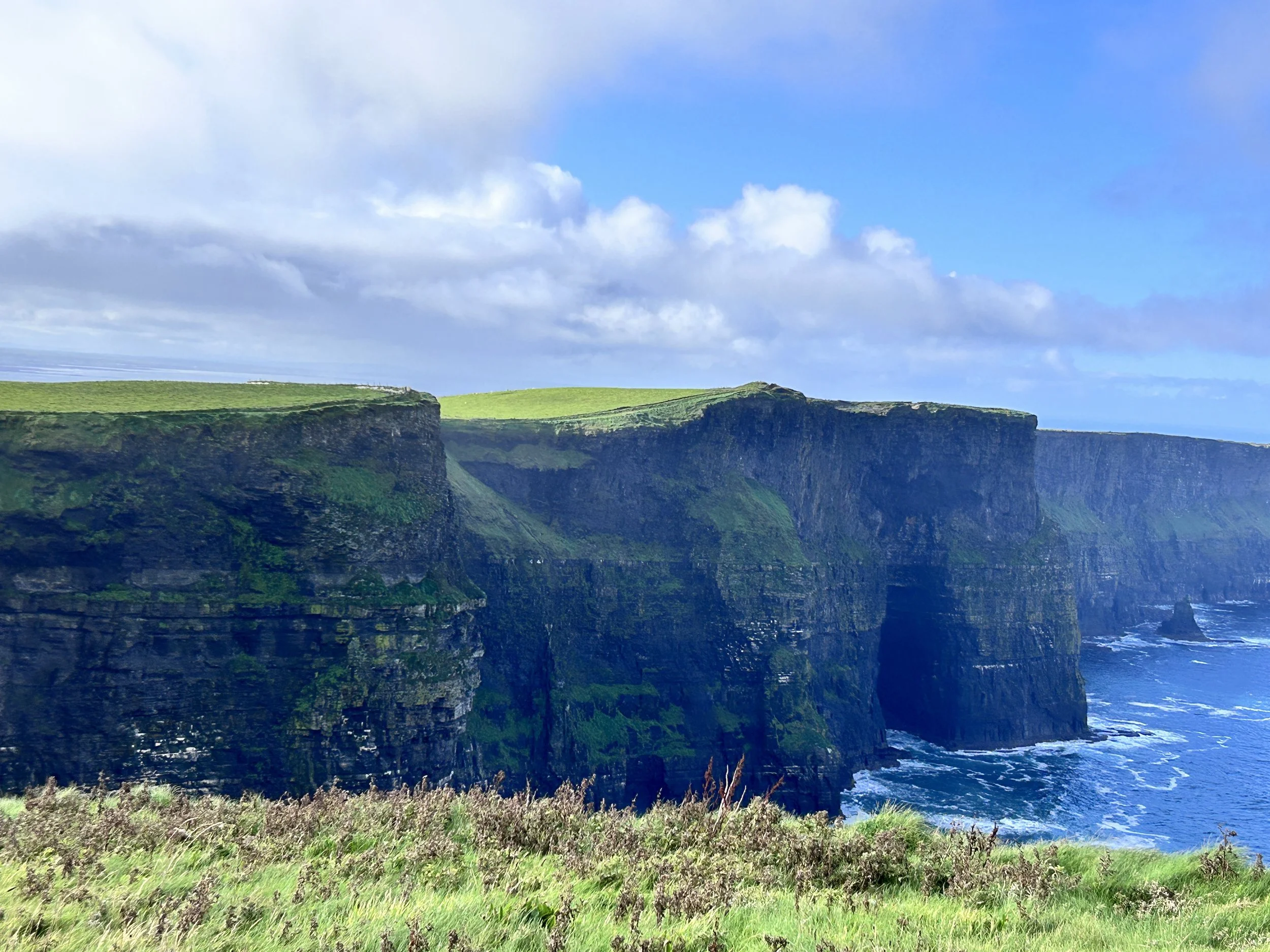 Cliffs with green grass on top, overlooking the ocean with waves hitting the rocks, under a partly cloudy sky in Ireland, Cliffs of Moher.