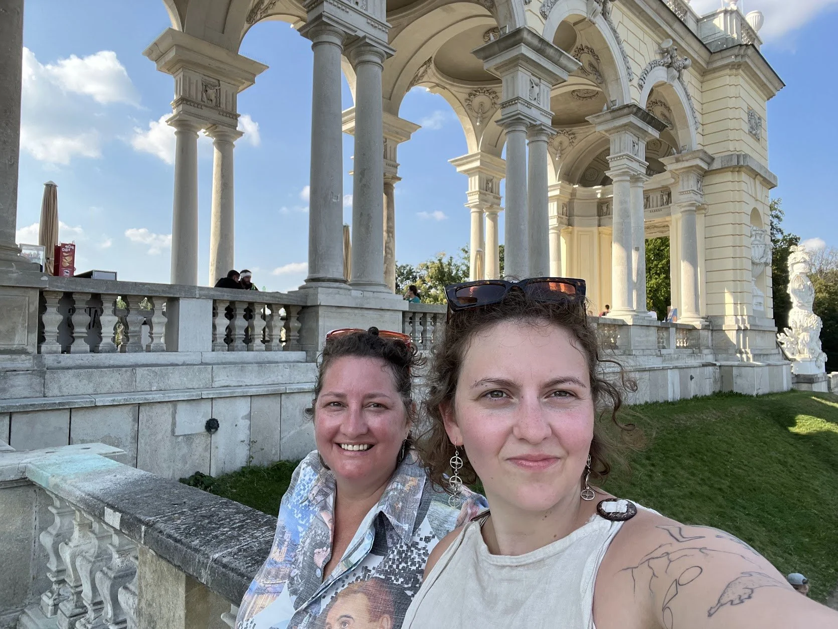 Two women smiling and taking a selfie in front of a white, ornate, columned architectural structure with a blue sky and scattered clouds in the background in the Schonnbrun gardens, Vienna Austria.