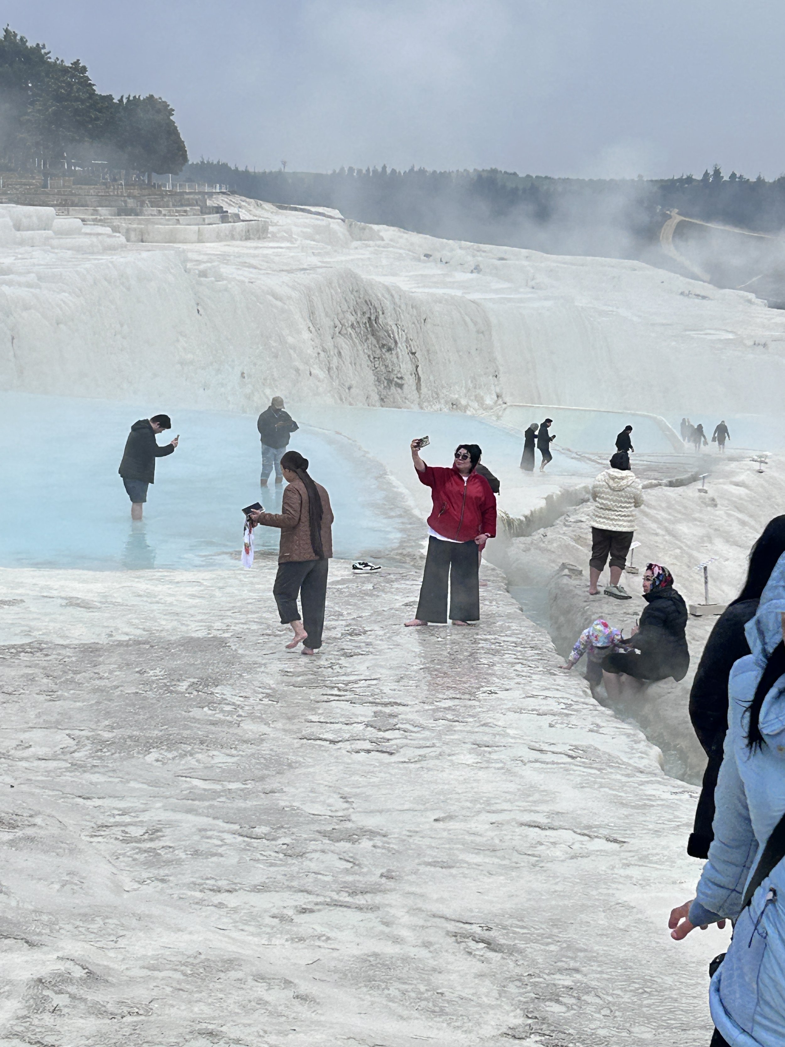 People visiting a hot spring or thermal pool at the travertine terraces of Pamukkale in Turkey, with some taking photos and others relaxing in the water.