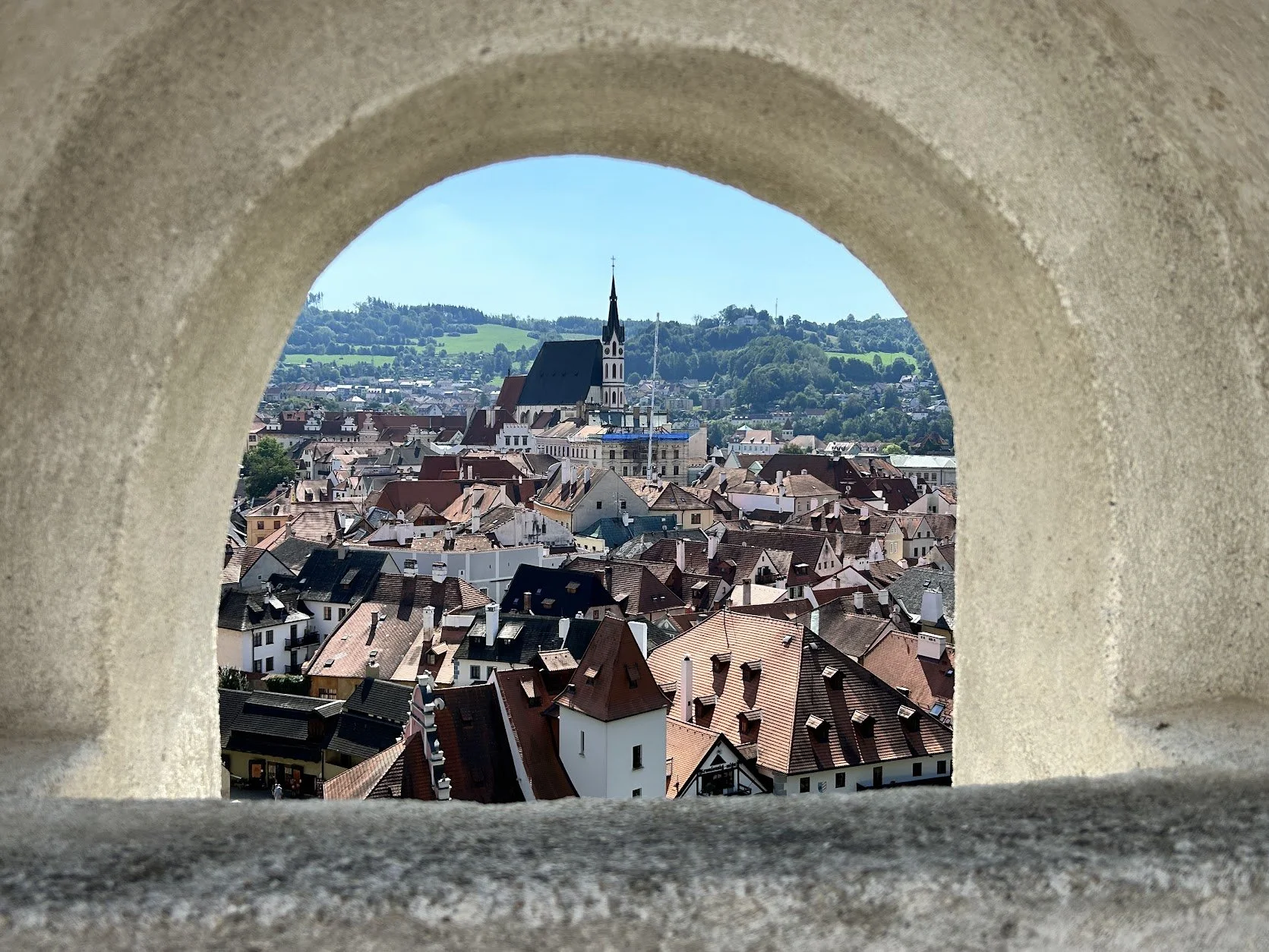 Cityscape viewed through a stone arch with red-tiled rooftops, a church with a tall steeple, and rolling green hills in the background in Cesky Krumlov.