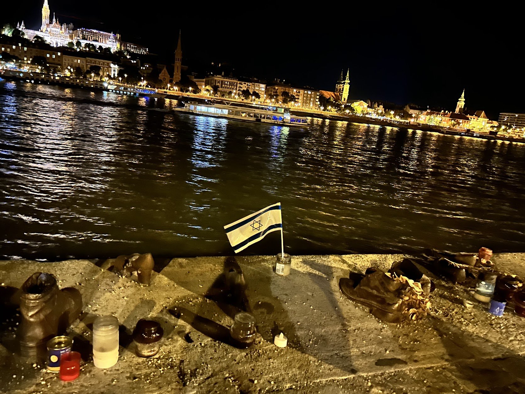 Nighttime scene by a river with illuminated cityscape in the background. Items on the ground include shoes, candles, and a small Israeli flag in a jar in Budapest.