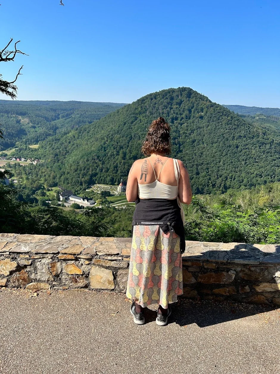 A woman stands with her back to the camera, overlooking a lush green valley and hills during daytime. View from an Abby in Austria.