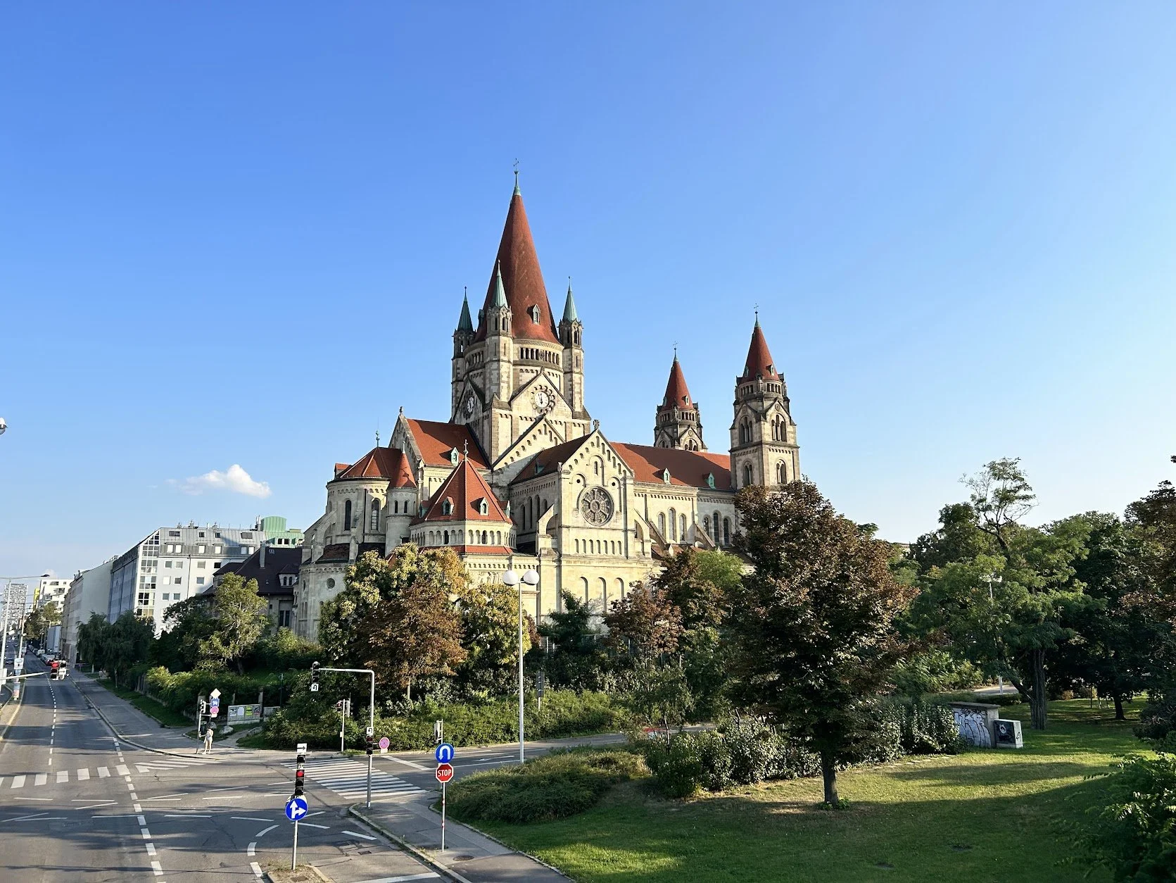 A large historic church with red-tiled spires and stone facade, surrounded by trees and a city street with traffic signs, under a clear blue sky in Vienna.
