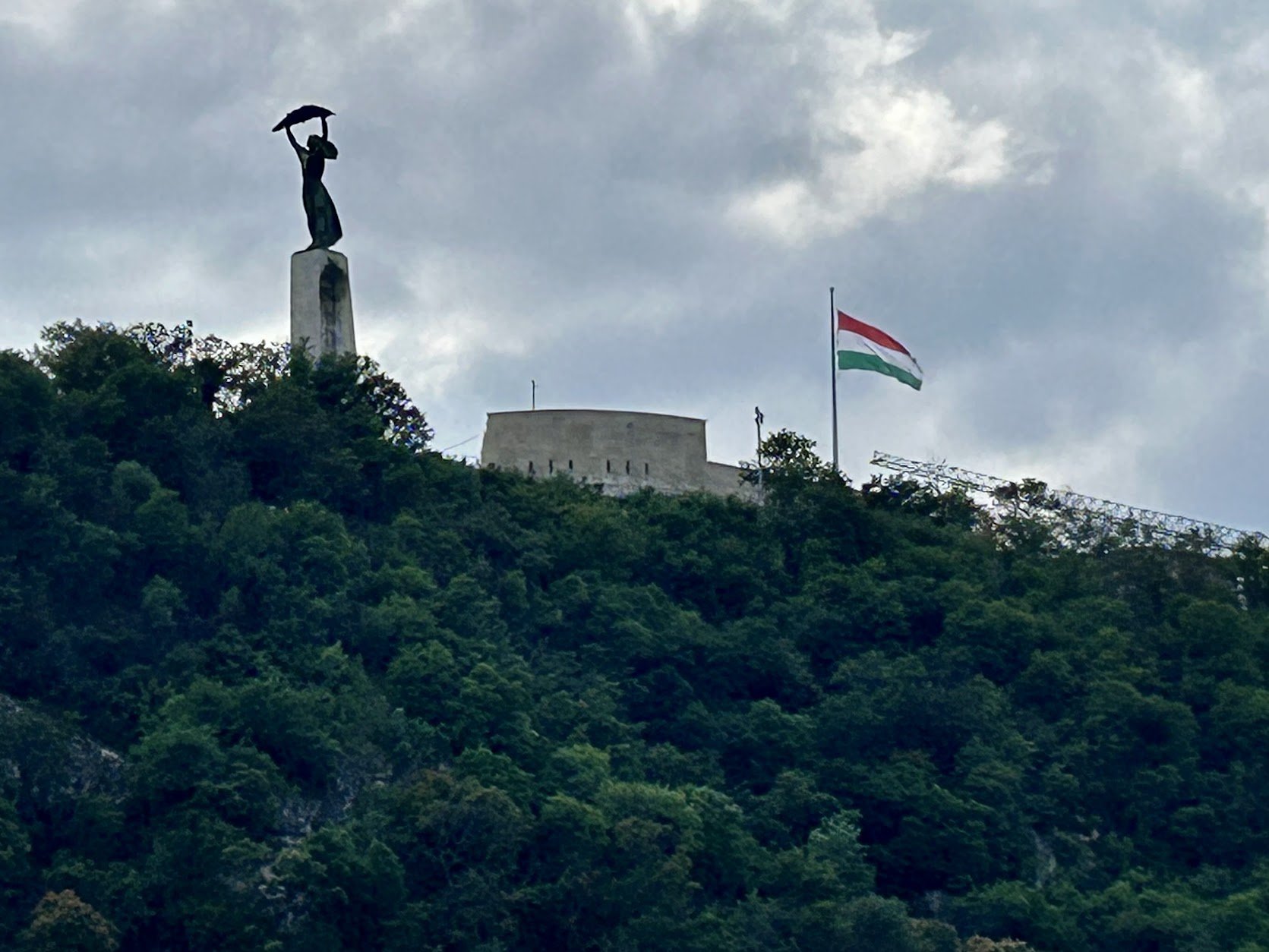 A hill covered with dense green trees features a statue of a woman holding a curved object above her head, standing on a tall pedestal. Freedom monument and Flag in Budapest.