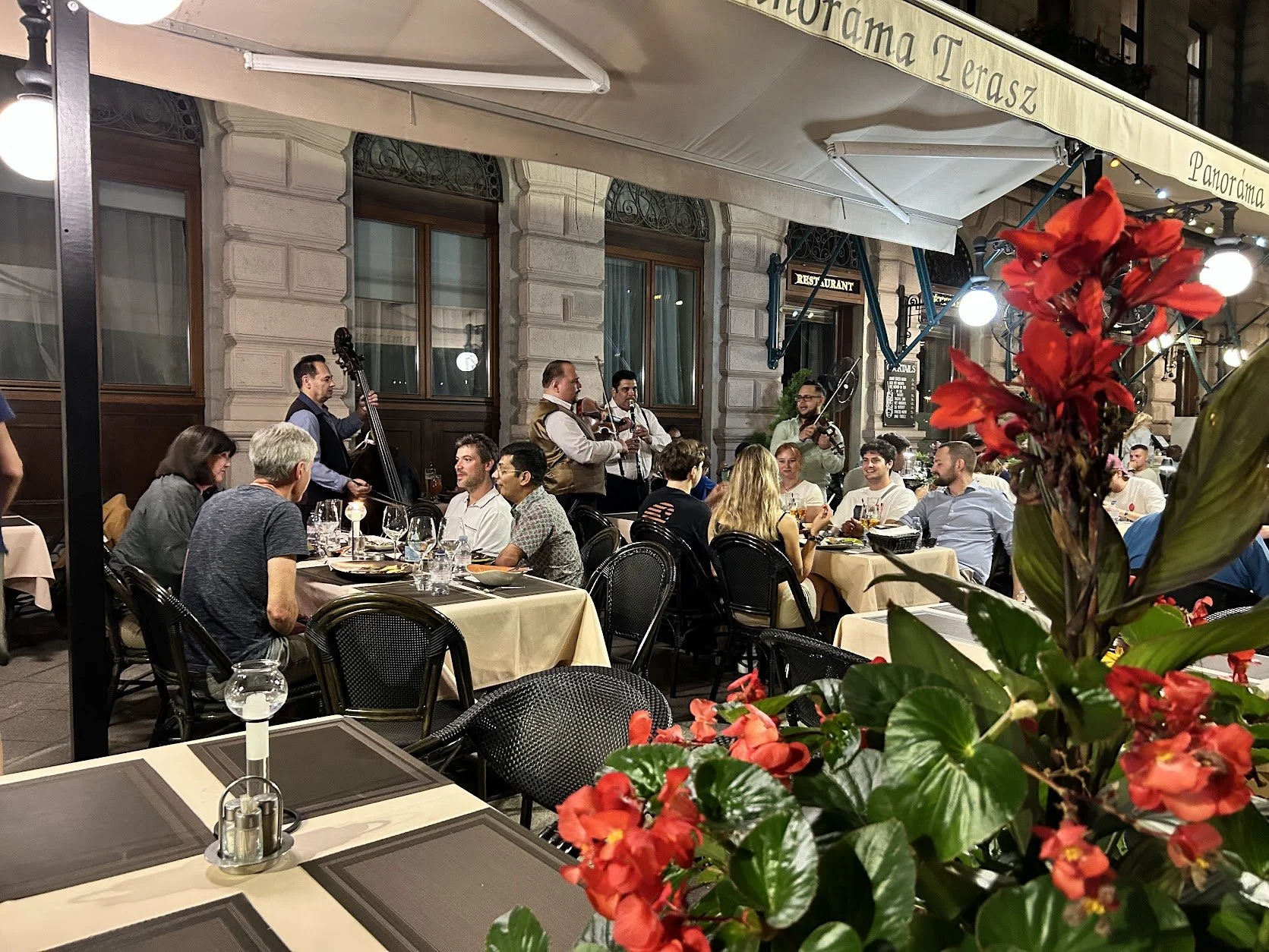 Outdoor restaurant at night with people dining and live jazz band playing music, decorated with red flowers in the foreground in Budapest.