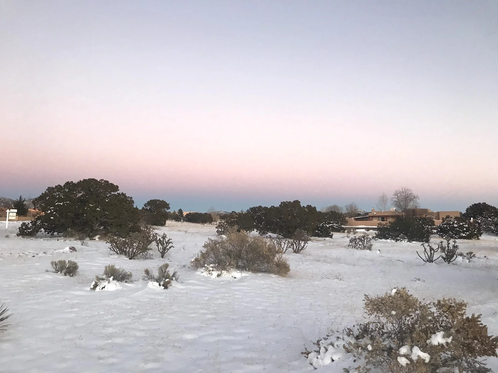 Snow-covered landscape with scattered bushes and trees, a house in the distance, and a colorful sky at dawn or dusk in New Mexico.