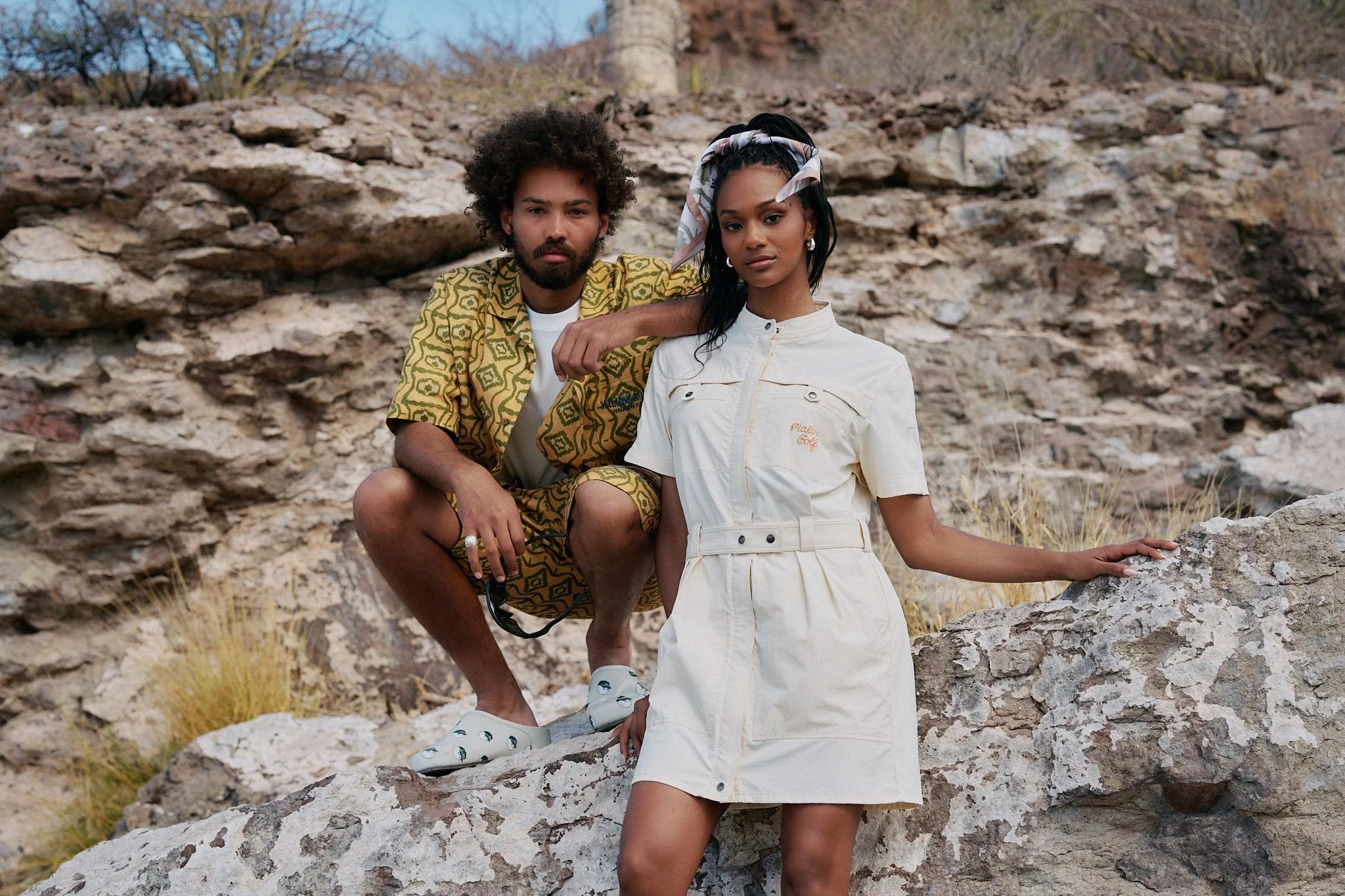 A man and woman posing on rocky terrain outdoors, with trees in the background. The man has curly hair and a beard, wearing a patterned yellow shirt. The woman has long black hair with a scarf tied around her head, wearing a cream-colored jumpsuit.