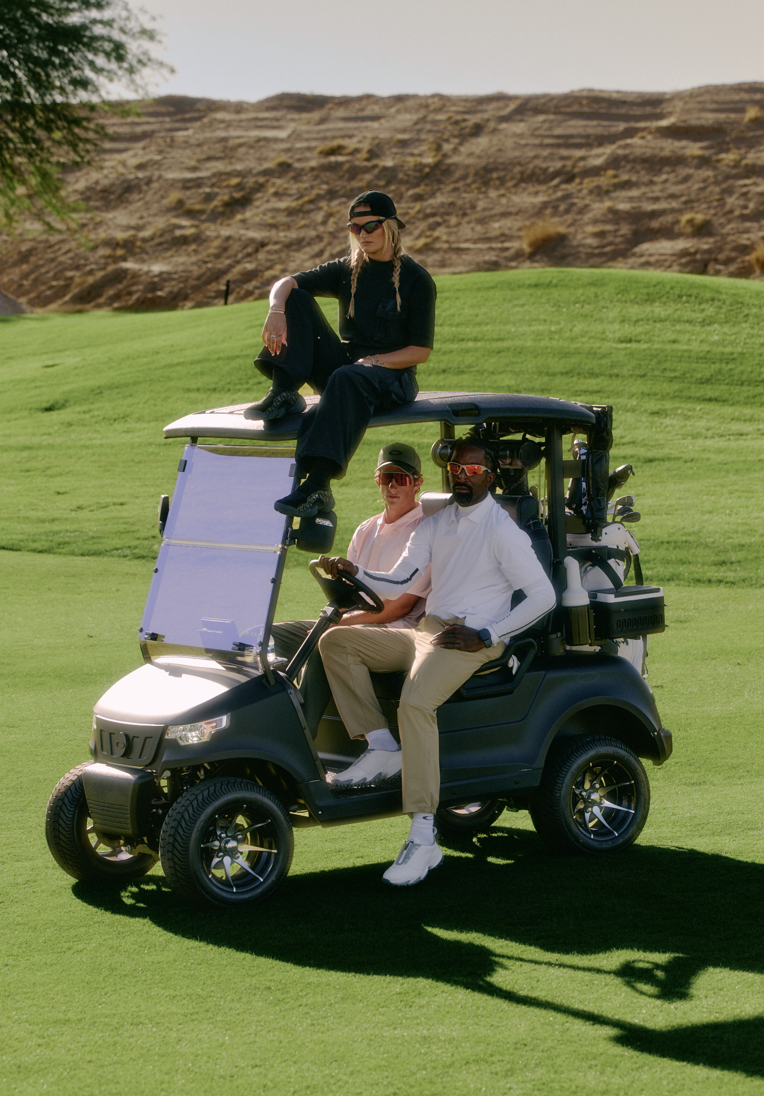 A golf cart with three people on a golf course. One woman is sitting on top of the cart wearing a black shirt, black pants, baseball cap backwards, sunglasses, and sporting braids. Inside the cart are two men, one wearing a white shirt and beige pant