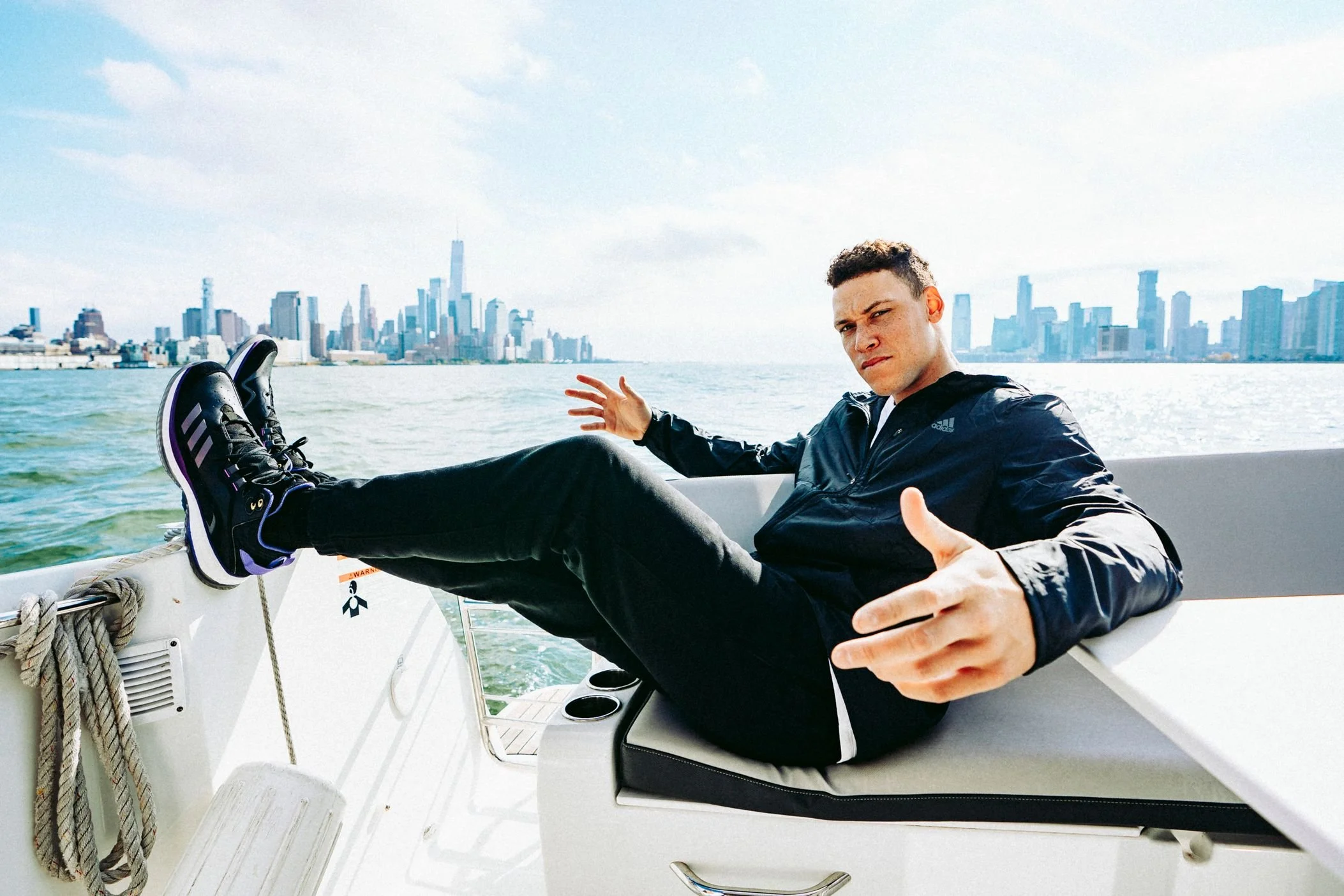 Young man relaxing on a boat, stretching out his leg with the New York City skyline in the background, including One World Trade Center, on a partly cloudy day.