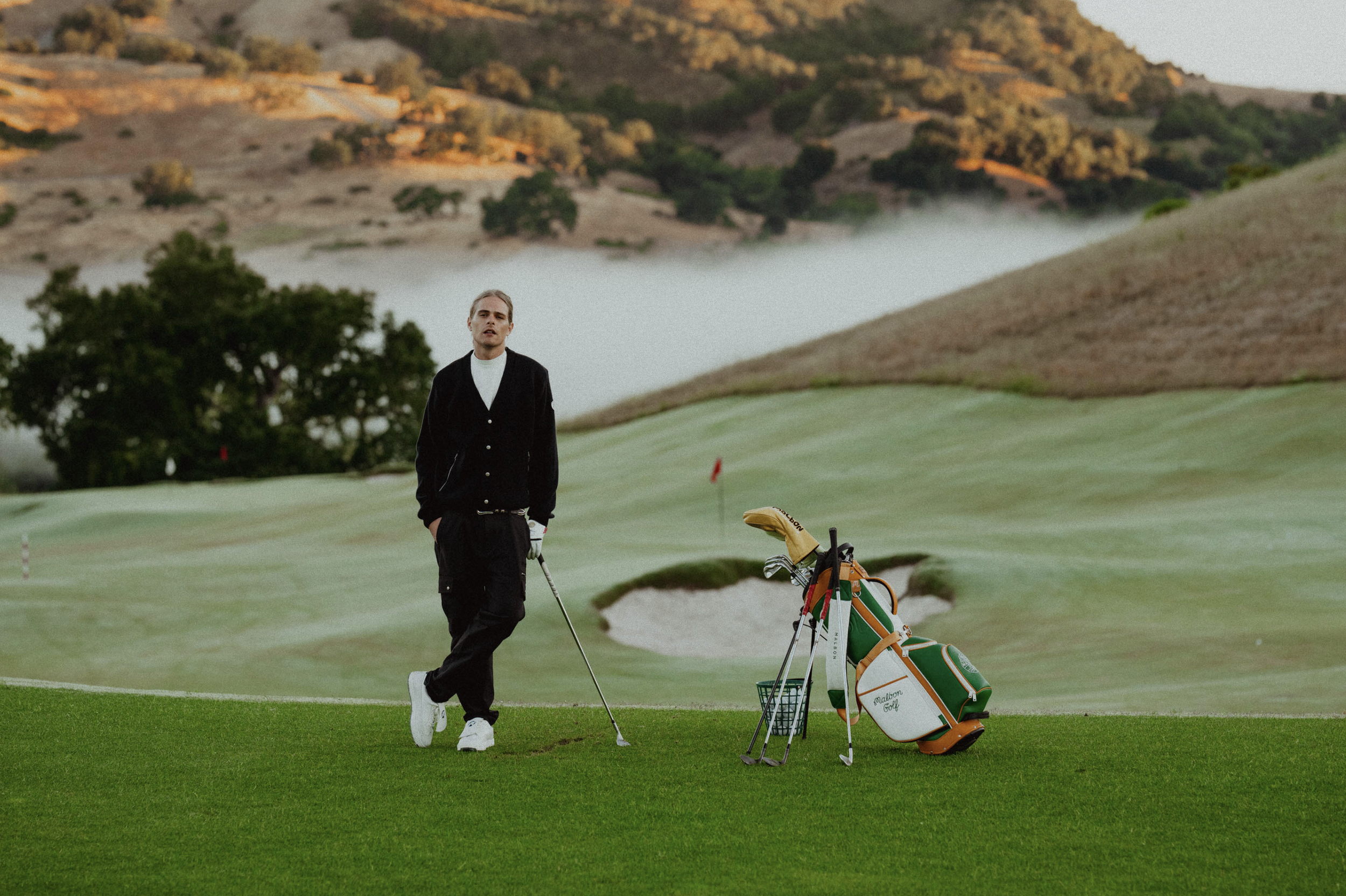 A man walking on a golf course holding a golf club, with a golf bag and cart nearby. The background features rolling hills and trees, with fog over the landscape.