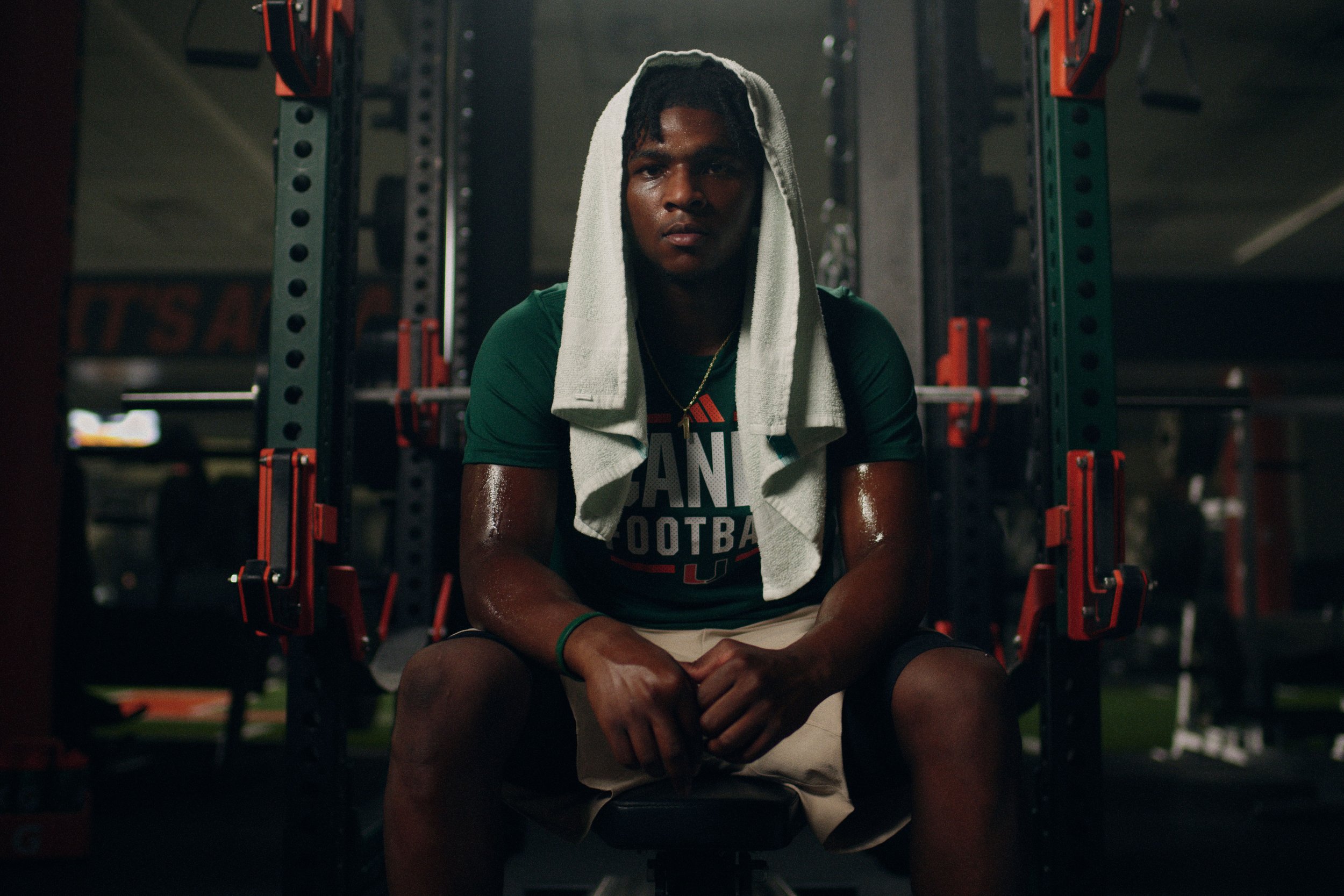 A young man sitting on a bench inside a weight room, wearing workout clothes and a towel over his head, appearing exhausted after exercising.