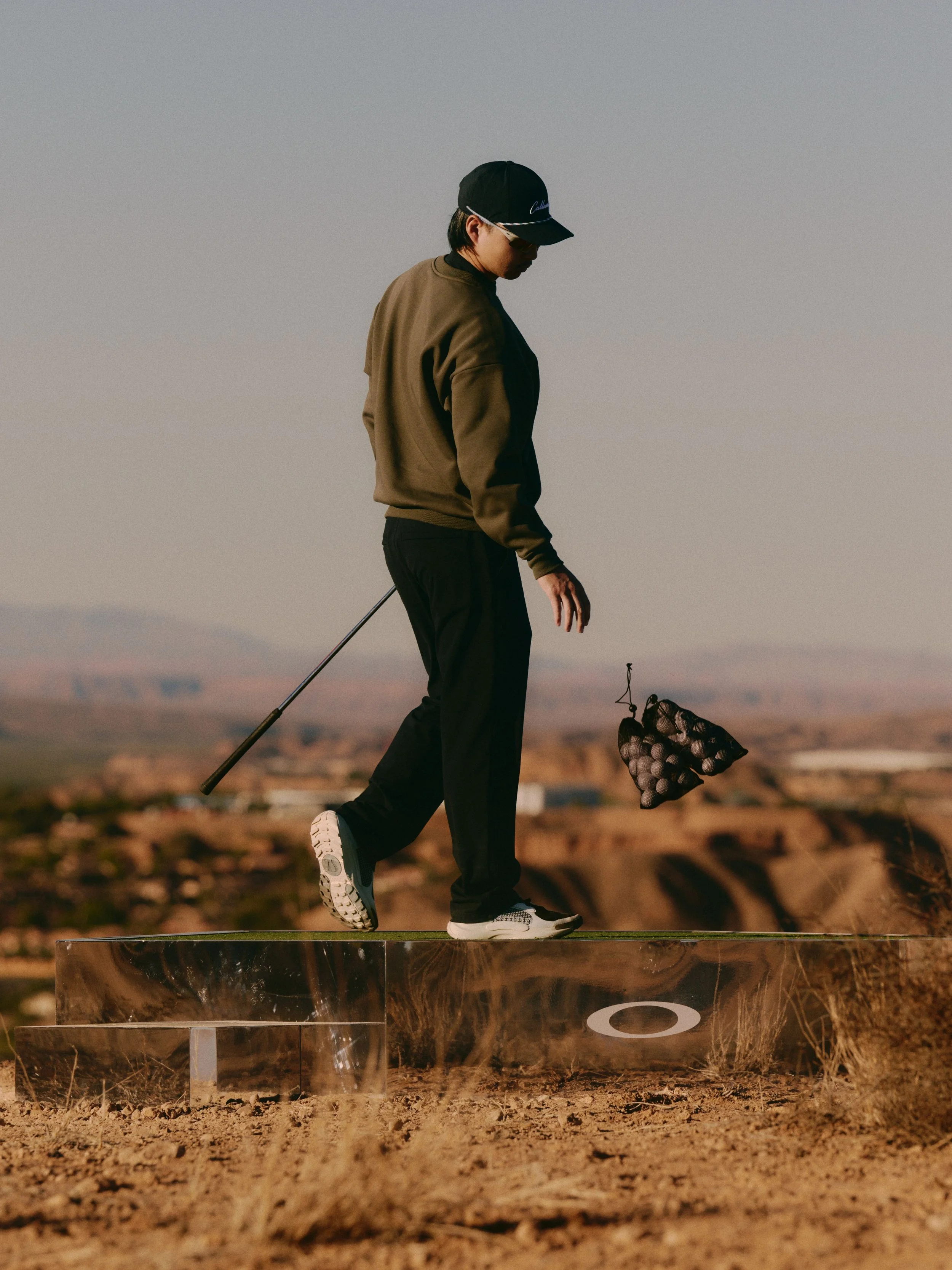 A person standing on a clear, transparent platform outdoors, wearing a black cap, brown jacket, black pants, and white sneakers, holding a bag of golf balls in one hand and a golf club in the other, with a vast landscape in the background during dusk
