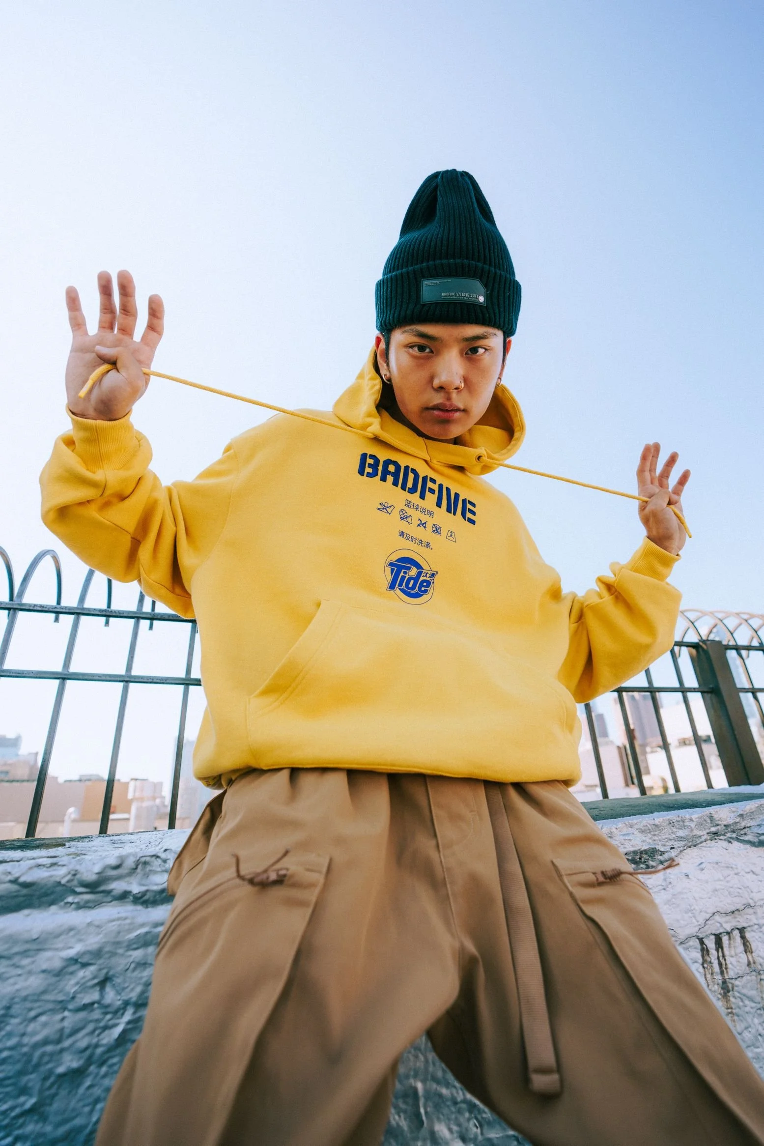 A person wearing a yellow hoodie with blue text and graphic, beige cargo pants, and a teal beanie, holding a yellow cord, standing outdoors near a metal fence, with a cityscape in the background, overcast sky.