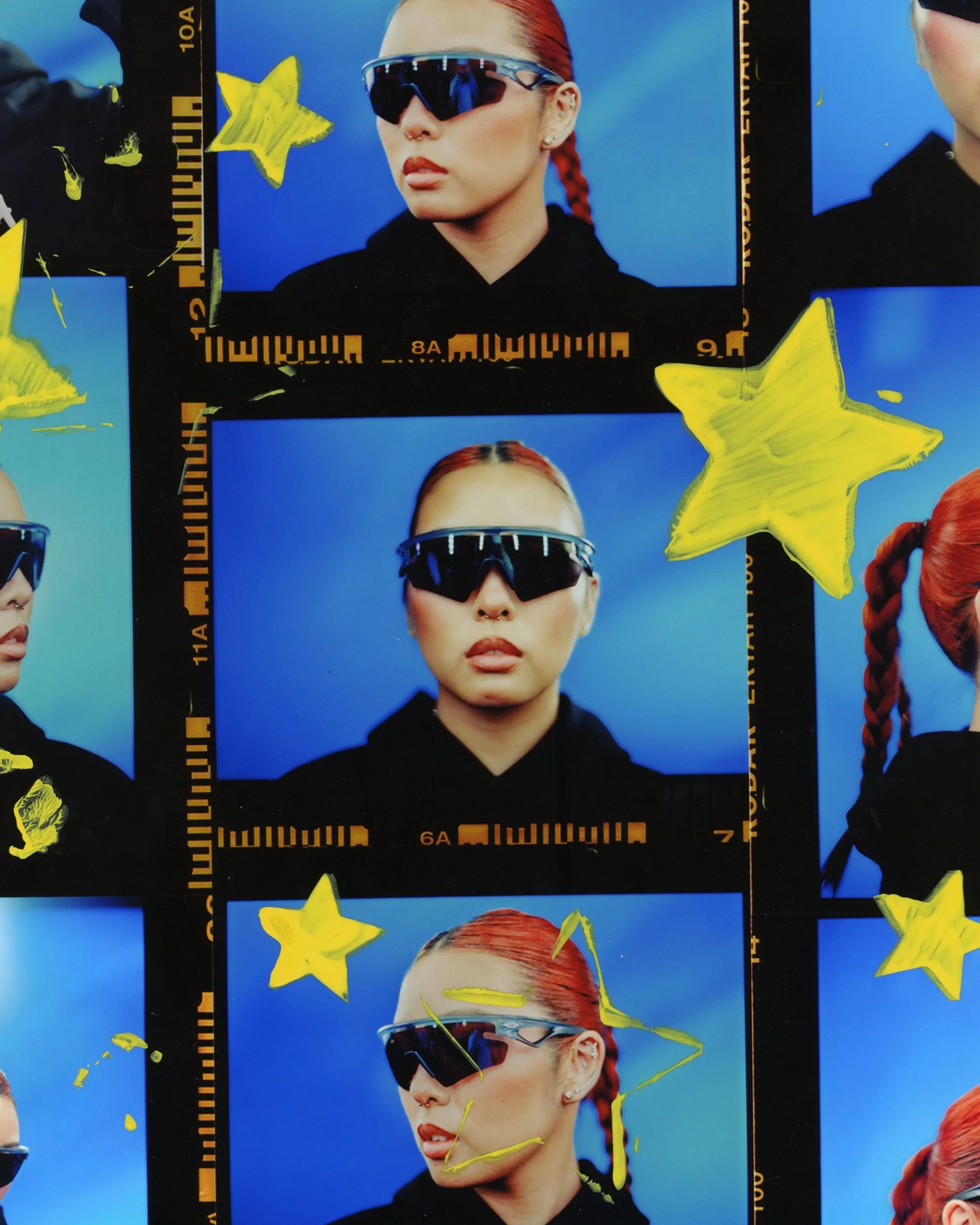 A close-up portrait of a woman with red hair tied back, wearing reflective sports sunglasses and a black top, set against a bright blue background with yellow star decals.