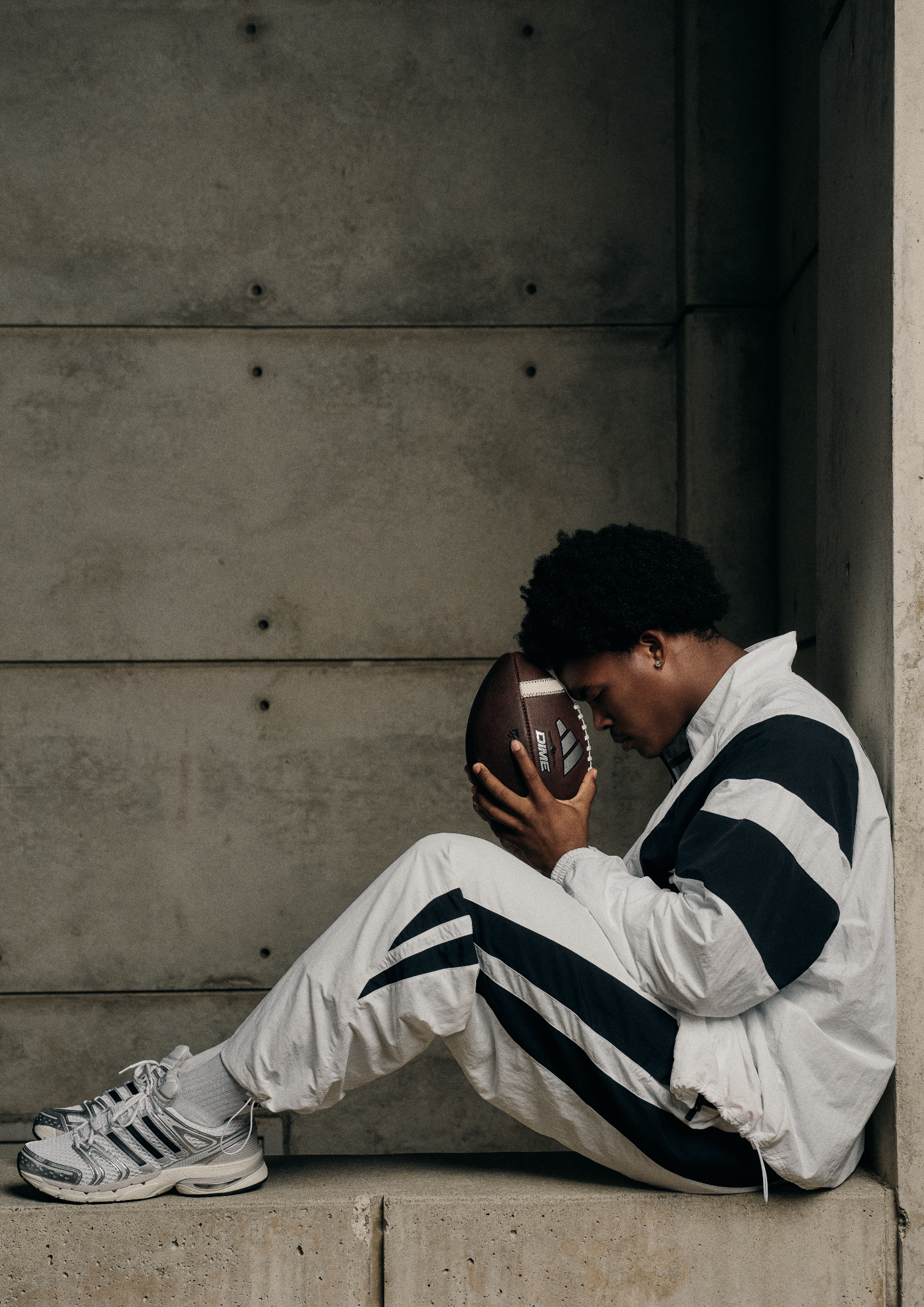 A young person with dark curly hair wearing a black and white tracksuit and sneakers, sitting on a concrete ledge with their head bowed, holding an American football close to their face against a concrete wall background.